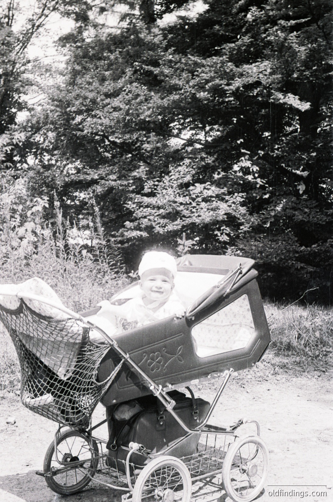 Vintage black-and-white photo of child in a vintage stroller with "SX" branding, set in a wooded area. The child wears a white cap and light clothing, suggesting mid-20th century (1950s–1960s).