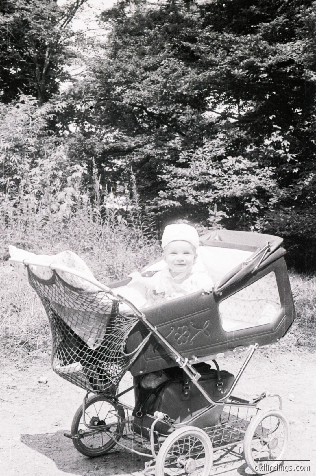 Vintage black-and-white photo of a child in a classic metal-framed pram on a forested path, mid-20th century. The pram’s basket detail and wooden wheels suggest mid-century design. Forest foliage and dirt path imply a rural or suburban setting.