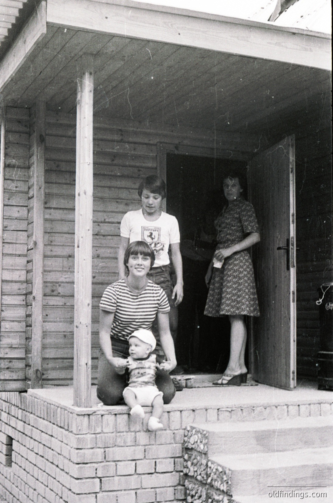 Family portrait on a brick porch, 1960s-1970s. Three individuals pose: a woman in floral dress holding a baby, a man in striped shirt, and a child in a white shirt with a Ferrari logo. Wooden house with simple wooden porch columns and a green door. Candid, nostalgic family moment.