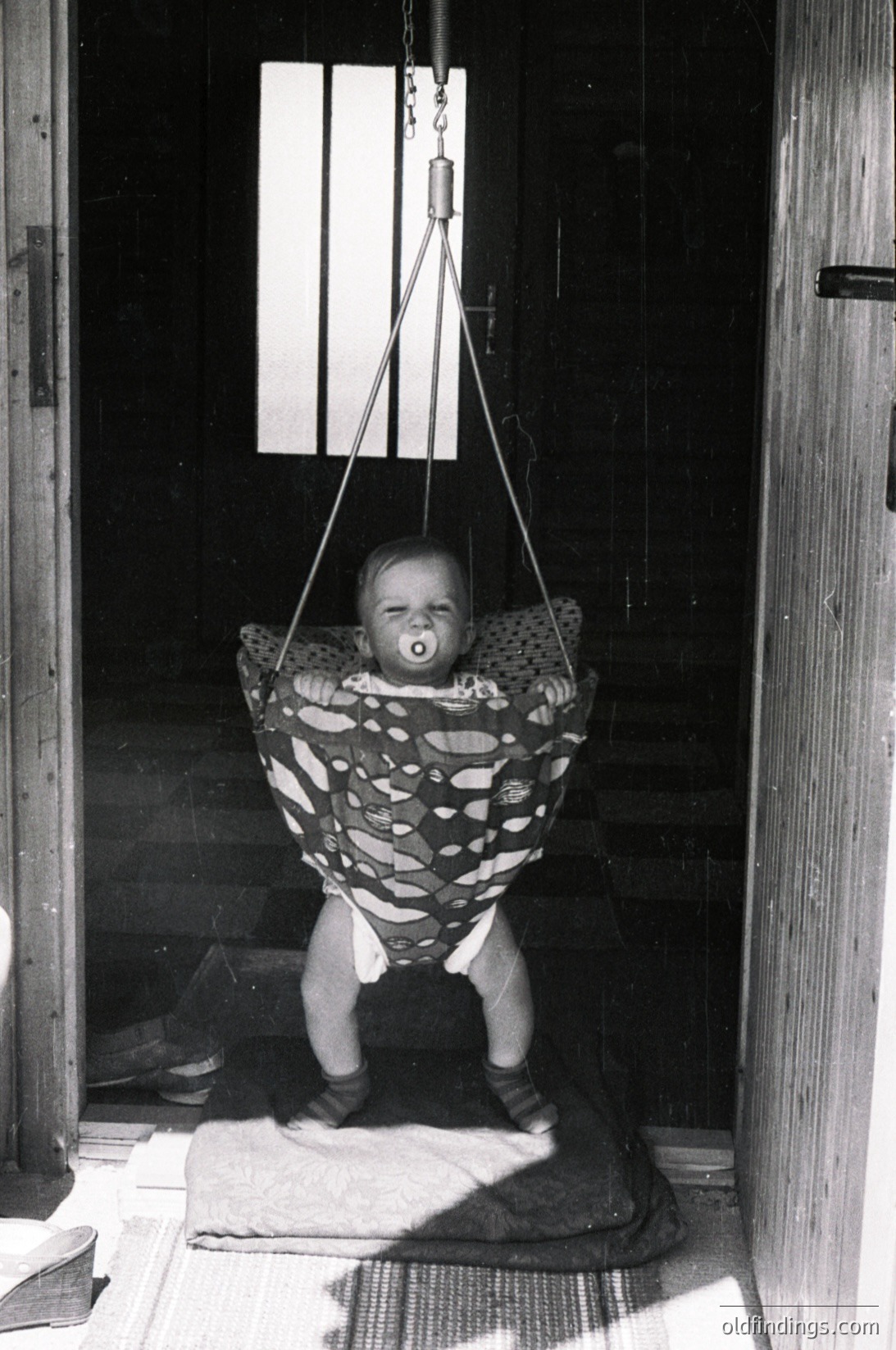 Vintage black-and-white photo of an infant suspended in a fabric swing, holding a pacifier, inside a rustic wooden doorway. Sunlight streams through the frame, illuminating the child’s patterned diaper. Mid-20th century domestic setting, likely 1950s–1960s.