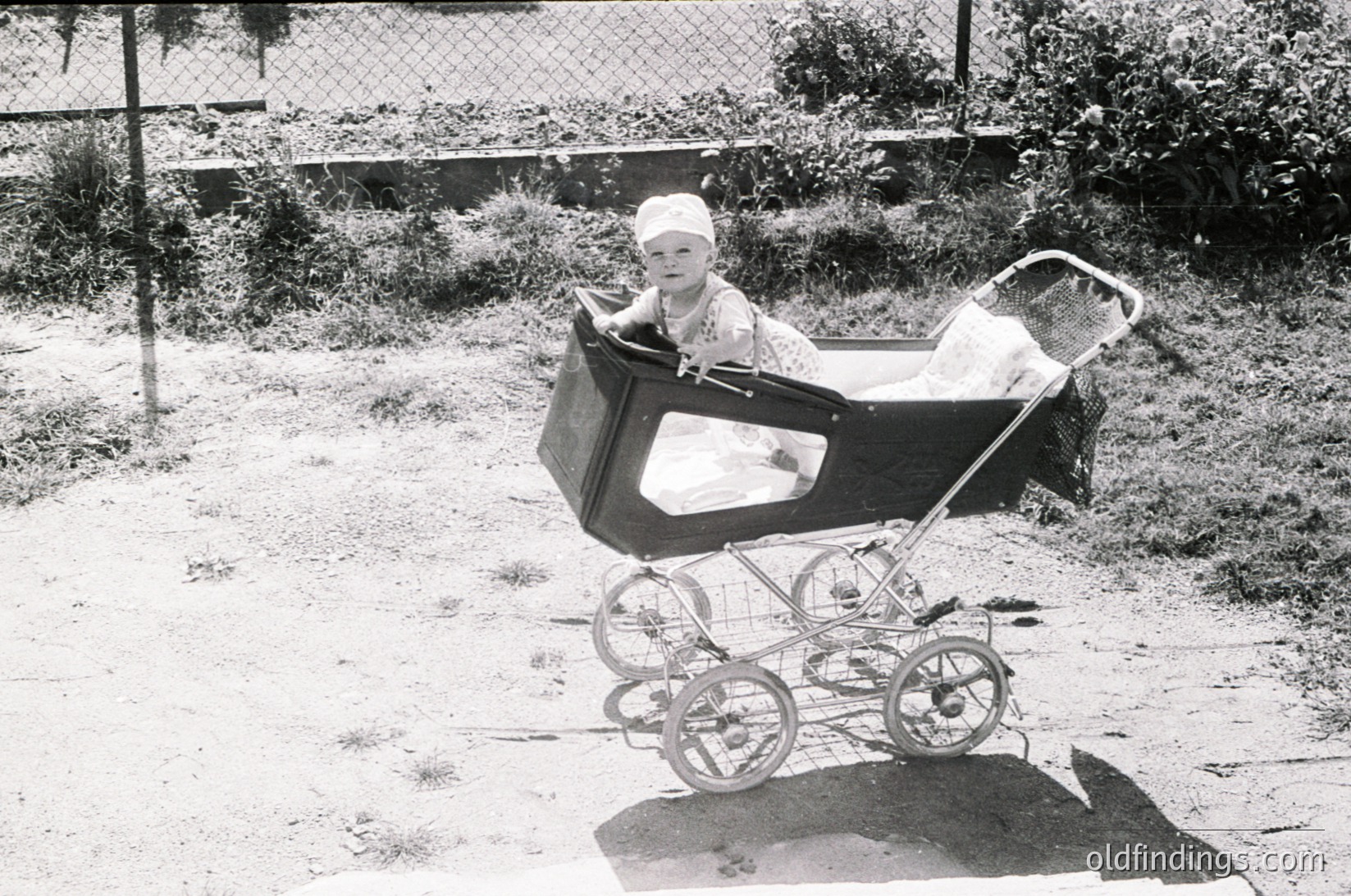 Vintage black-and-white photo of a child seated in a classic wicker pram with wire-spoked wheels, mid-20th century. Overgrown backyard with chain-link fence and brick wall in background. Infant wears a white bonnet and striped outfit, holding a folded cloth.