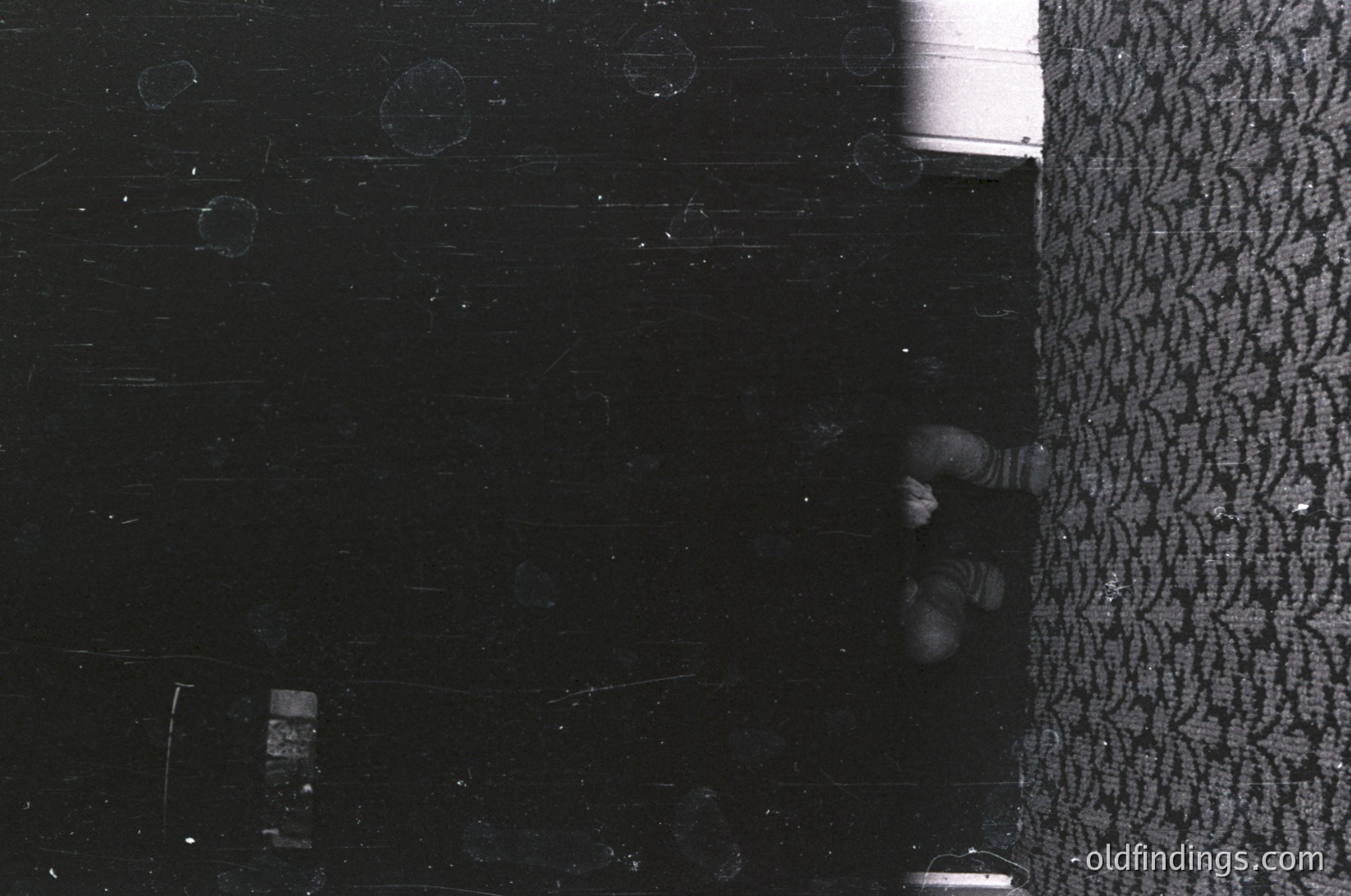 Vintage black-and-white photo of a child peeking from behind a patterned curtain, likely 1950s–1970s. Interior shows worn linoleum flooring with scuff marks and a small object (possibly a toy or book) on the floor. Candid, intimate snapshot of childhood curiosity.