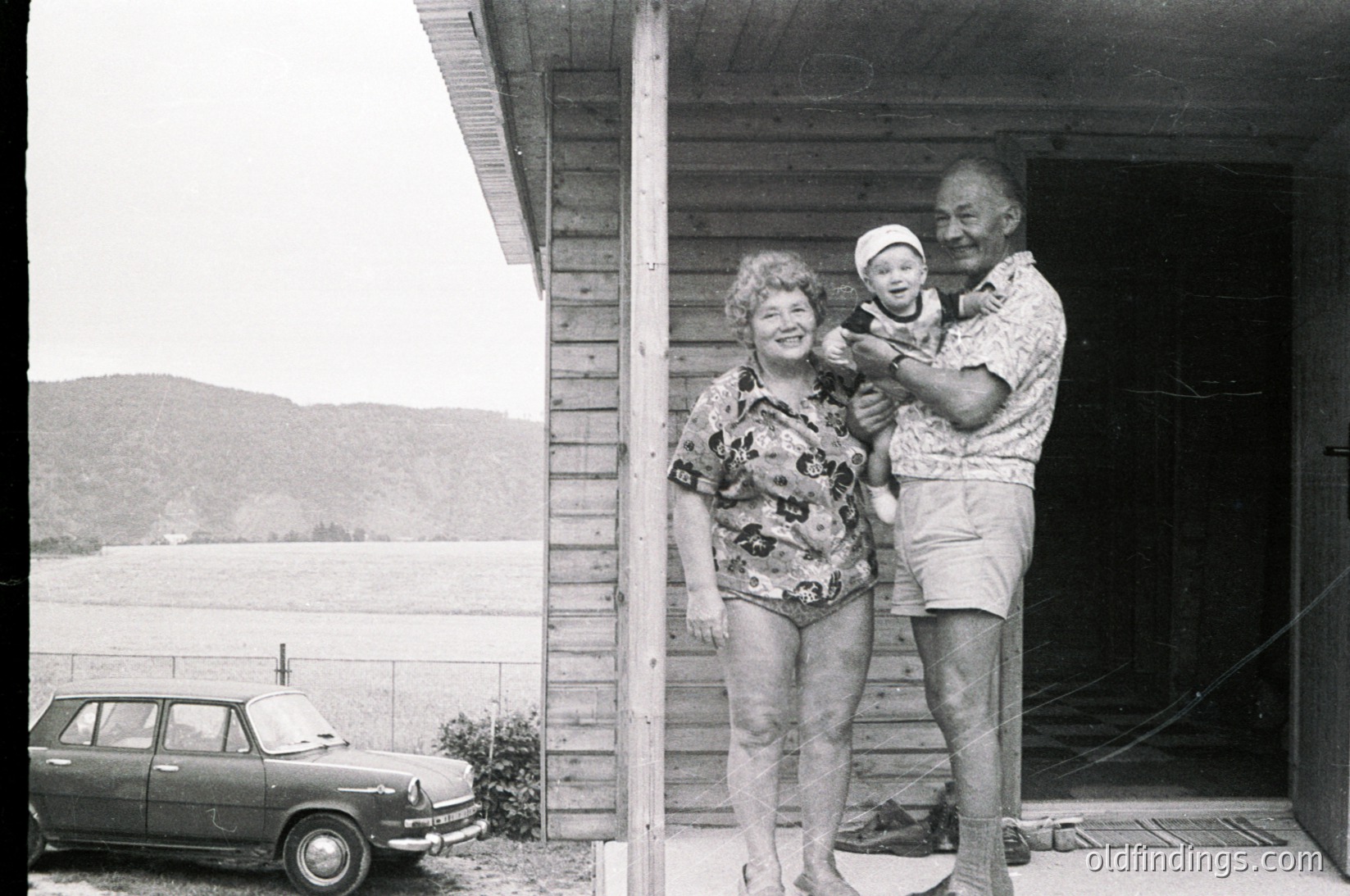 A mid-20th-century family portrait by a lakeside wooden cabin. Elderly couple (man in patterned shorts, woman in floral swimsuit) pose with a baby in a doorway. Classic vintage car (likely ) parked beside them. Mountain backdrop suggests alpine or forested lake region. Likely –.