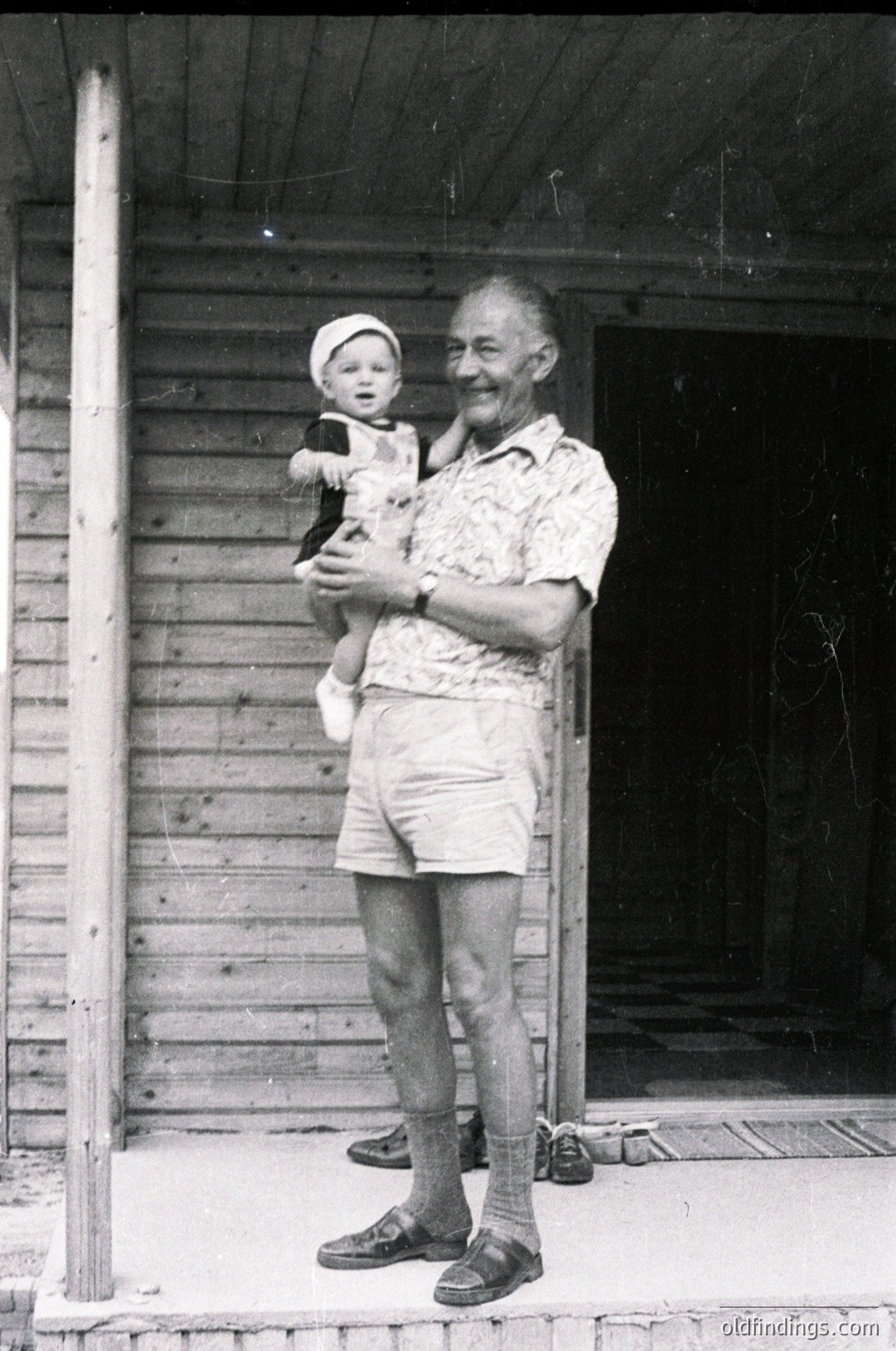 A mid-20th-century black-and-white photo of an elderly man holding a toddler on a wooden porch. The man wears a patterned shirt, knee-high socks, and sandals; the child wears a hat and a light dress. Rustic wooden plank construction and a simple ceiling fan suggest a rural or small-town setting, likely mid-1900s.
