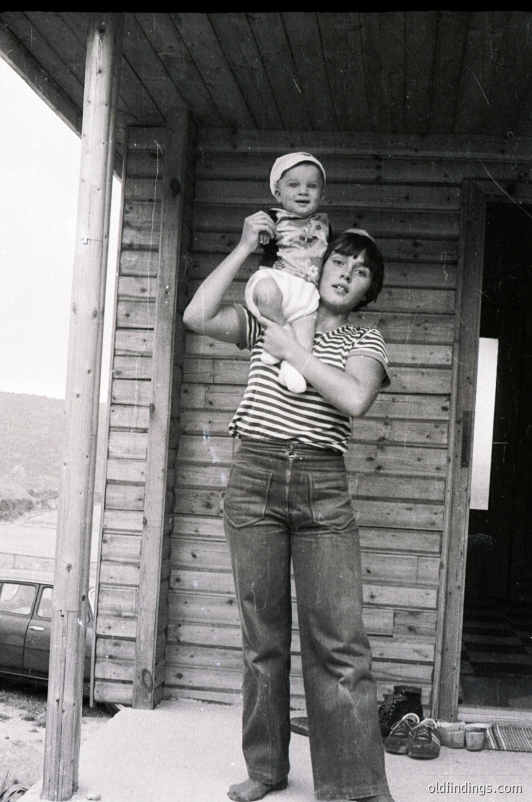 Black-and-white candid of a woman cradling a young child outside a rustic wooden structure, likely a rural home. She wears a striped shirt and wide-leg pants, while the child sports a knitted cap. The setting suggests mid-20th century (1950s–1970s) in a farming or village community.