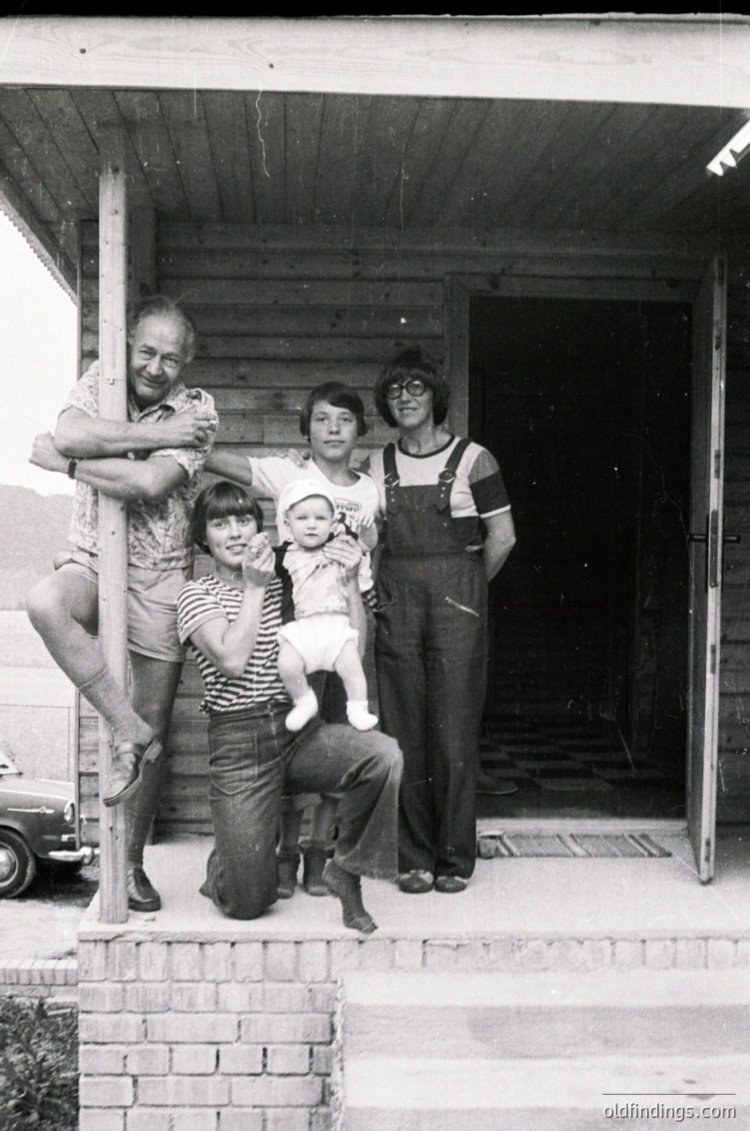 Family portrait in front of a rustic wooden structure, likely a barn or farmhouse porch, dated to the 1970s. Five individuals pose: an elderly man, two adults in overalls, a woman in a striped dress, and a baby. The setting suggests rural life, with brick steps and a vintage truck in the background.