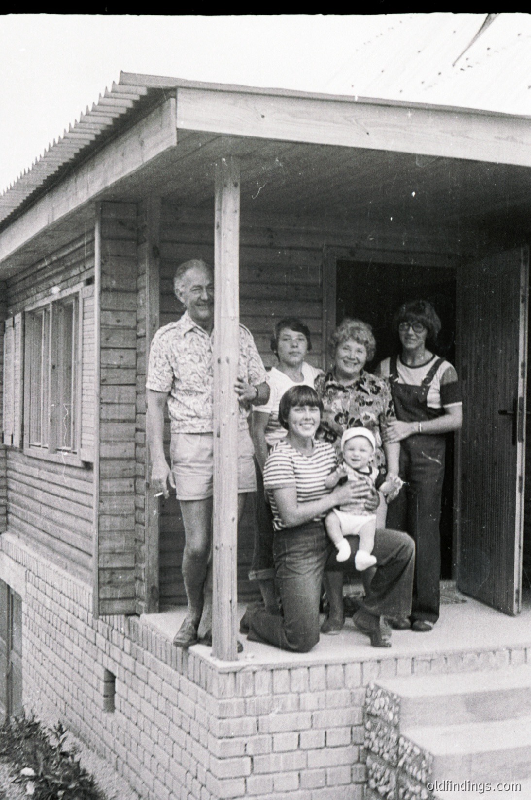 Family portrait in front of a wooden house, likely from the 1970s–1980s. Five adults and a child pose on a brick porch with a simple wooden overhang. Striped and patterned clothing, overalls, and a baby carrier suggest mid-century European suburban life.