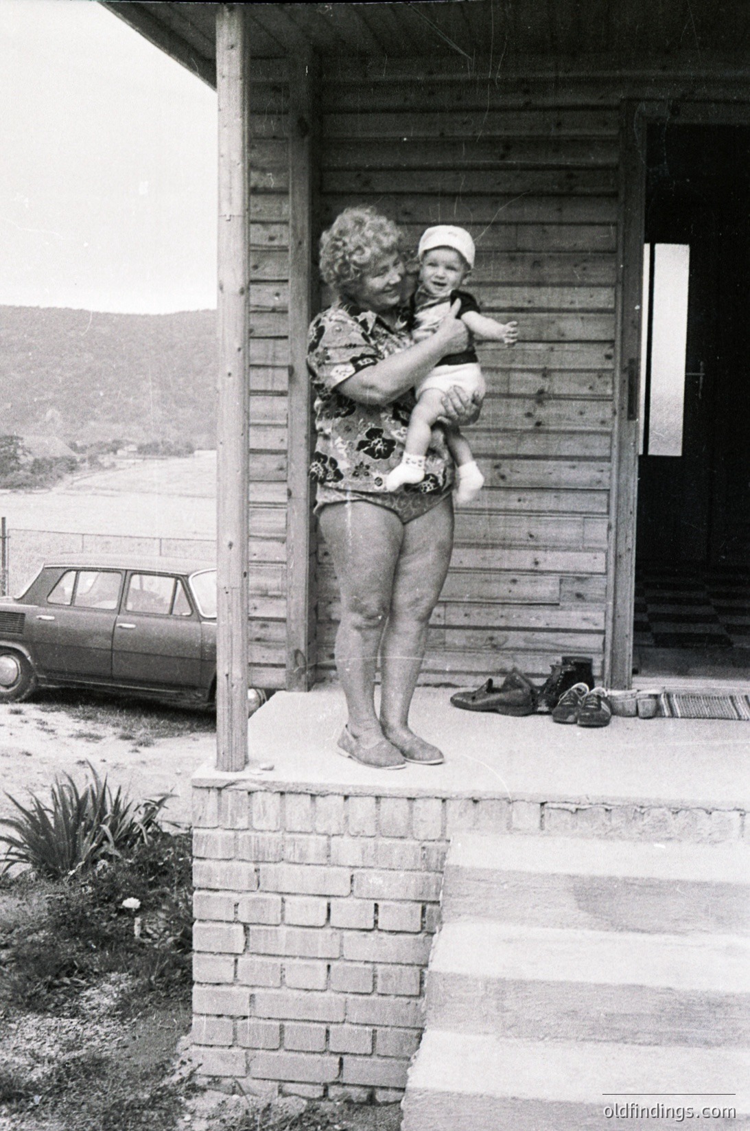 A woman in a floral swimsuit cradles a baby in a white cap on a wooden balcony, overlooking a coastal road. Classic car parked below, vintage shoes discarded on steps. Likely 1960s–1970s seaside setting.