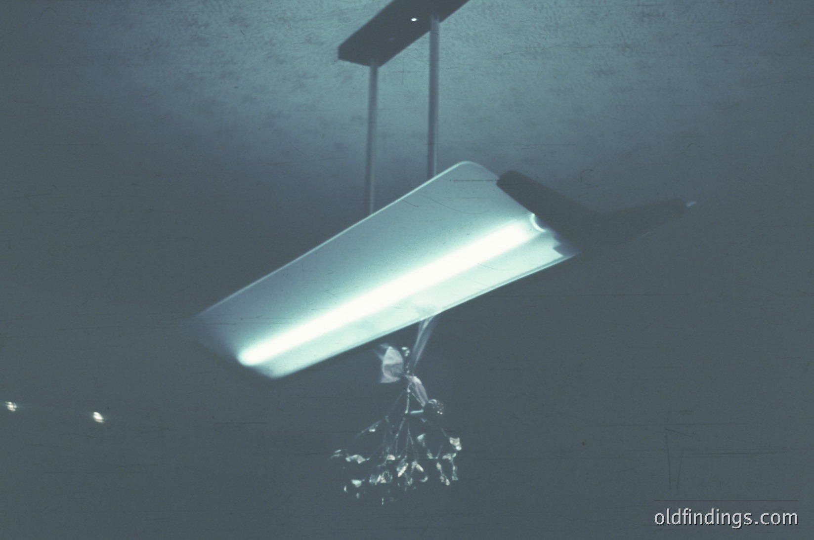 Industrial-style fluorescent light fixture suspended from a rough concrete ceiling, paired with a minimalist crystal chandelier. Moody lighting creates dramatic contrast. Ideal for modern loft, gallery, or vintage-inspired interior design.