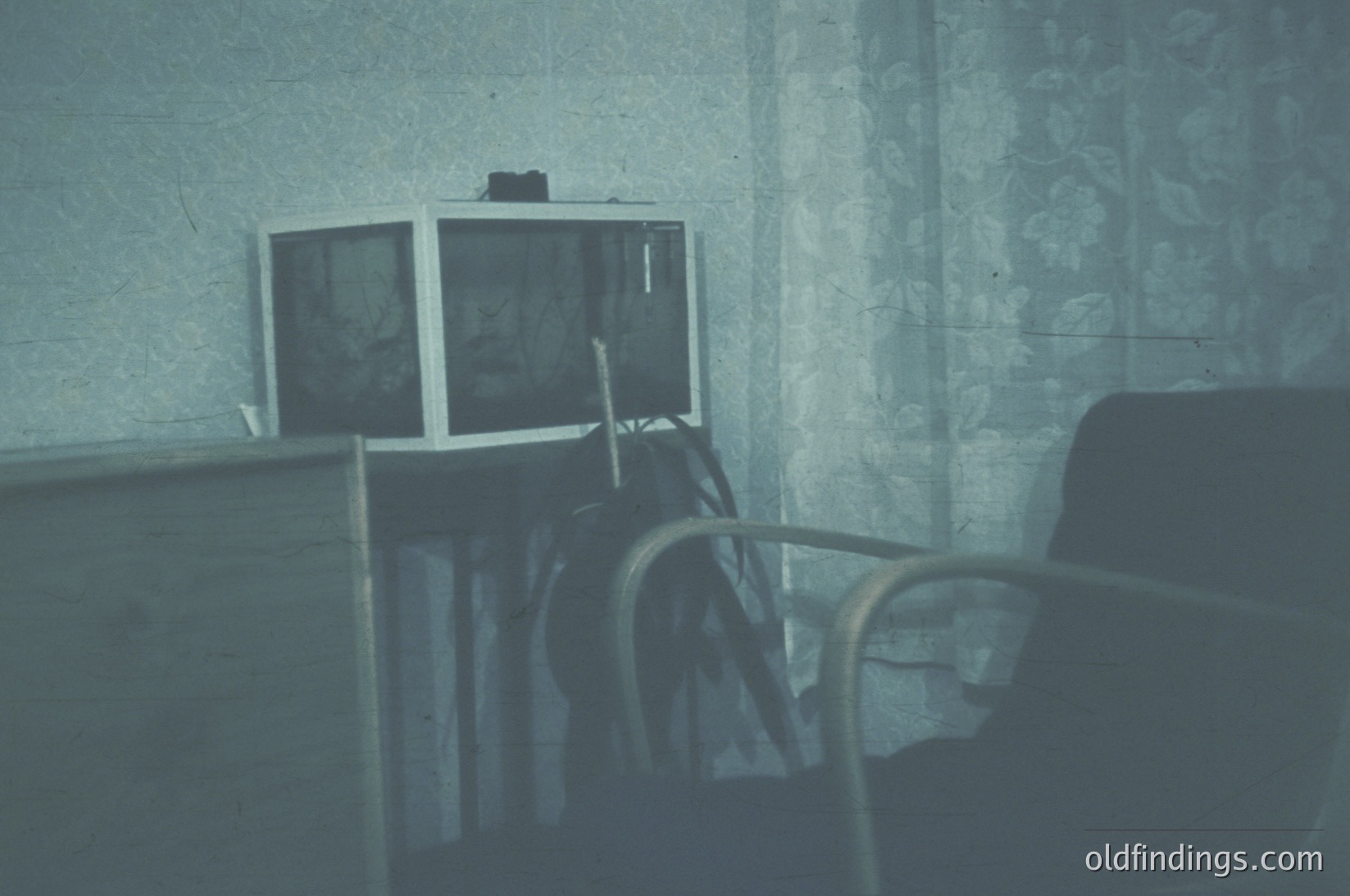 Vintage interior featuring a double-monitor setup on a wooden desk, likely from the 1970s–1980s. Monochrome CRT screens display faint text/images, flanked by a black office chair with metal framing. Floral-patterned wallpaper and worn linoleum floor suggest a mid-century office or educational setting.