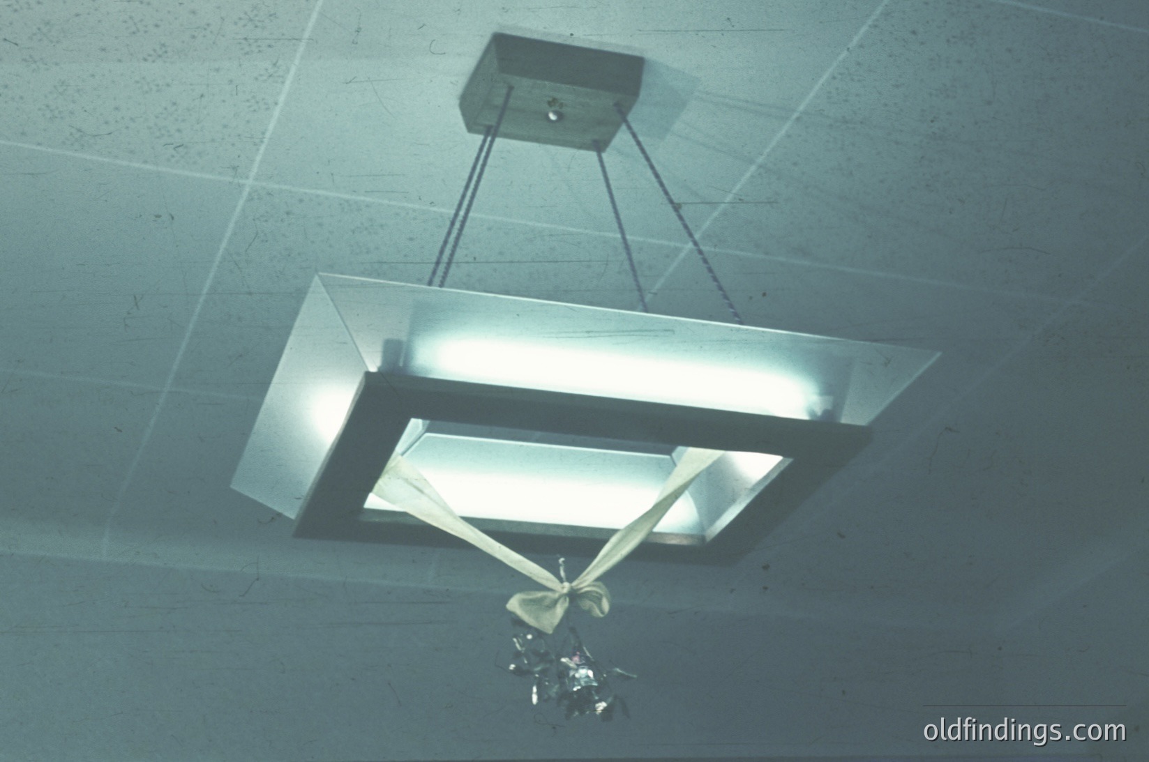 Minimalist industrial pendant light with rectangular fluorescent tube and geometric metal frame suspended from rough concrete ceiling. Decorative metallic ornament hangs below.