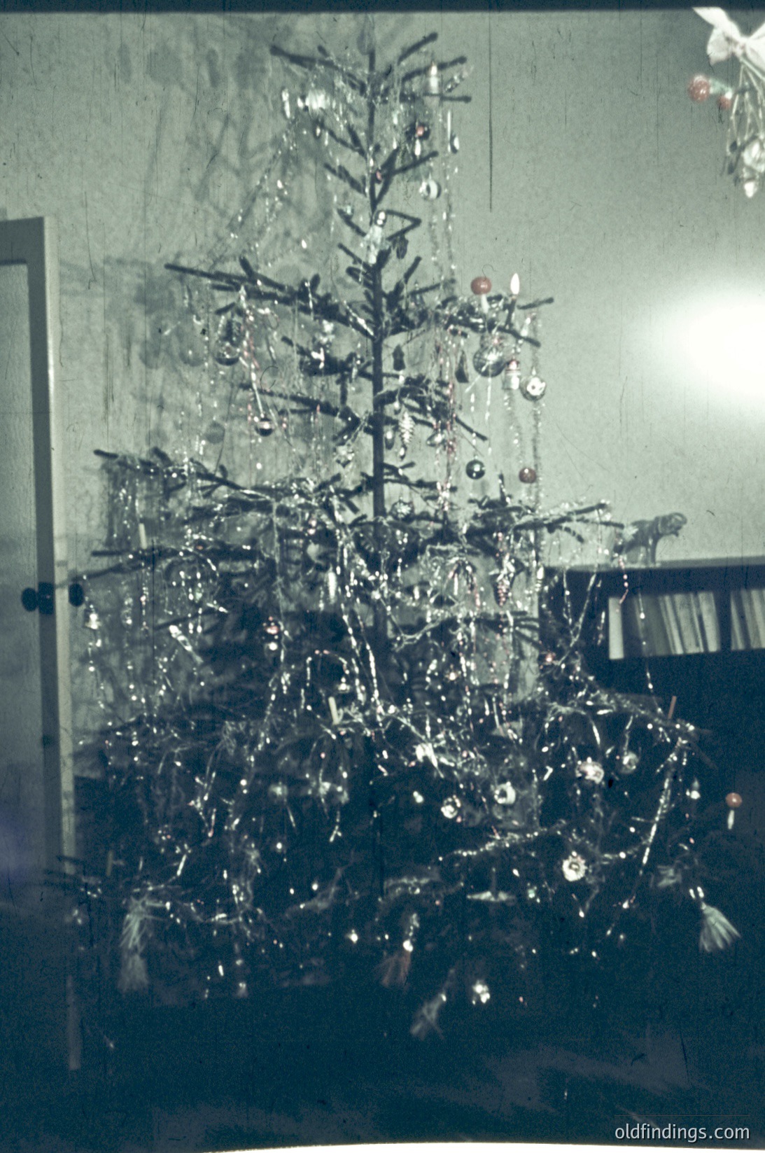Vintage Christmas tree adorned with metallic ornaments, tinsel, and a single red star topper in a modest indoor setting, likely mid-20th century. Plain walls and wooden furniture suggest a modest home decor style.