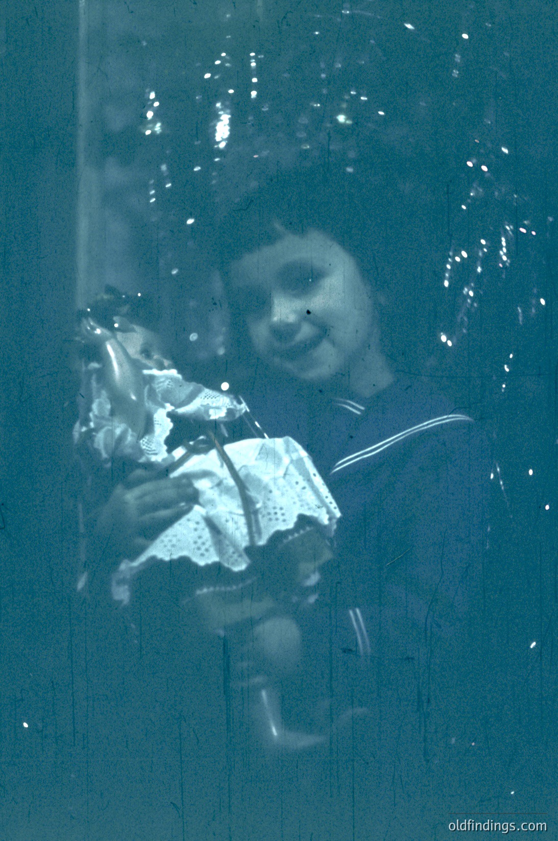 Vintage sepia-toned photo of a child peering through rain-streaked glass, holding a white lace handkerchief. Mid-20th century urban setting suggested by clothing and window framing. Evokes nostalgia and candid childhood moments.