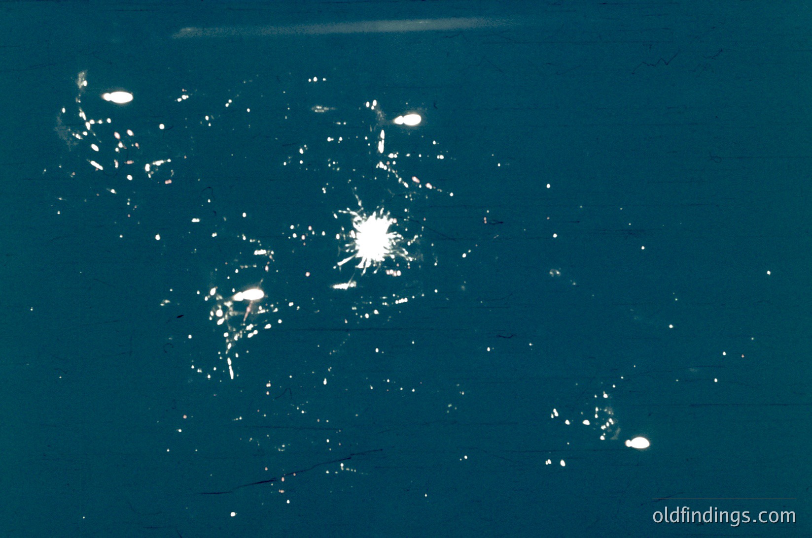 Aerial nighttime view of urban lights forming geometric patterns. Likely a satellite or drone capture showing city infrastructure and development density. The brightest areas suggest concentrated activity, possibly commercial or industrial zones. The scattered lights indicate residential or less dense regions.