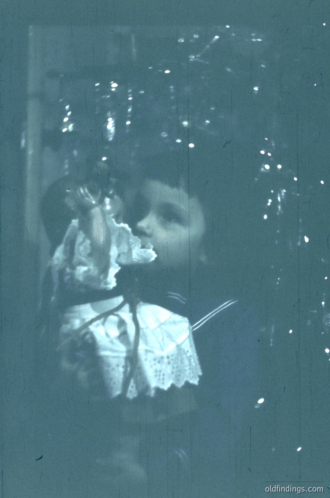 Vintage sepia-toned photo of a child in early 20th-century attire, holding a small object (likely a toy or tool) near their mouth. Blurred background suggests motion or focus on subject. Possible candid street or urban setting.