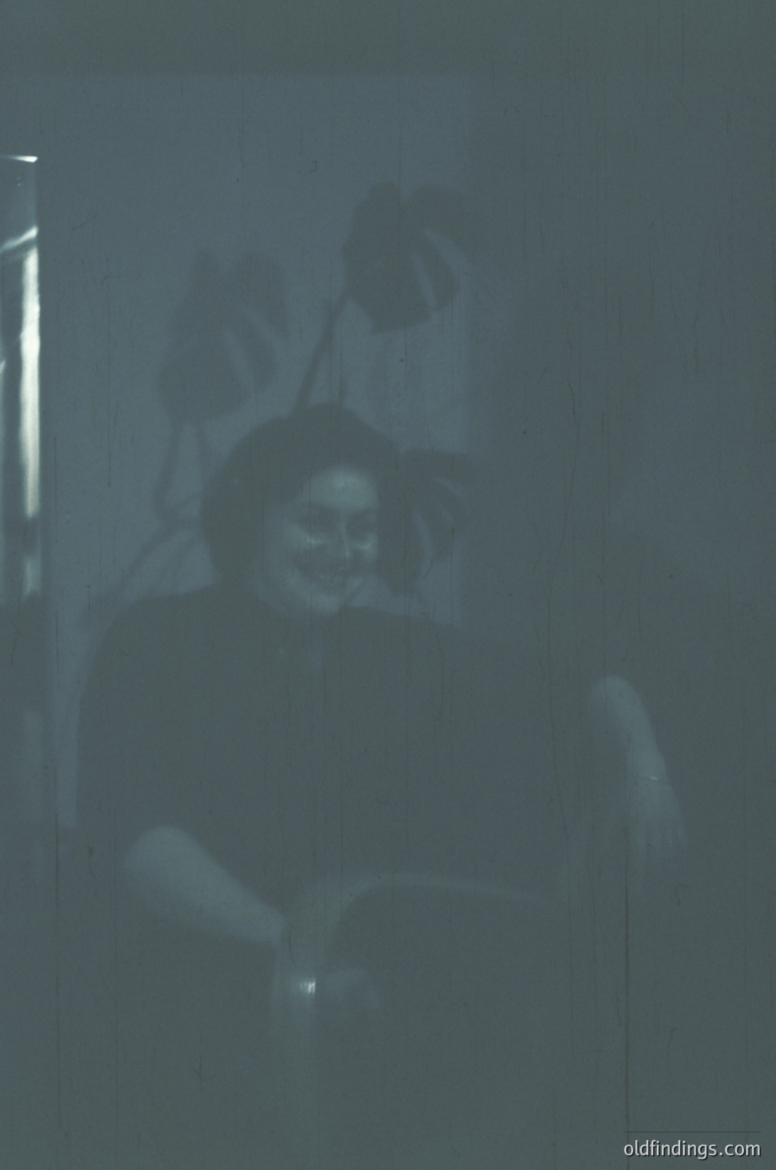 Vintage monochrome portrait of a seated individual in dim lighting, likely from the 1950s–1970s. The person holds a book or object, with a faintly visible lamp or light source behind them. Soft focus and low contrast enhance nostalgic, candid atmosphere.