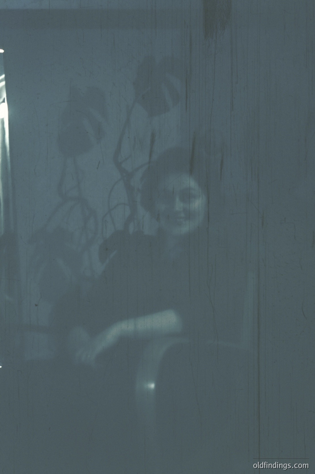 Silhouetted figure seated on a window ledge, back to camera, framed by a frosted glass pane. Urban setting with faint streetlight glow. Likely mid-20th century due to architectural style and lighting. Evokes mood of solitude or contemplation.