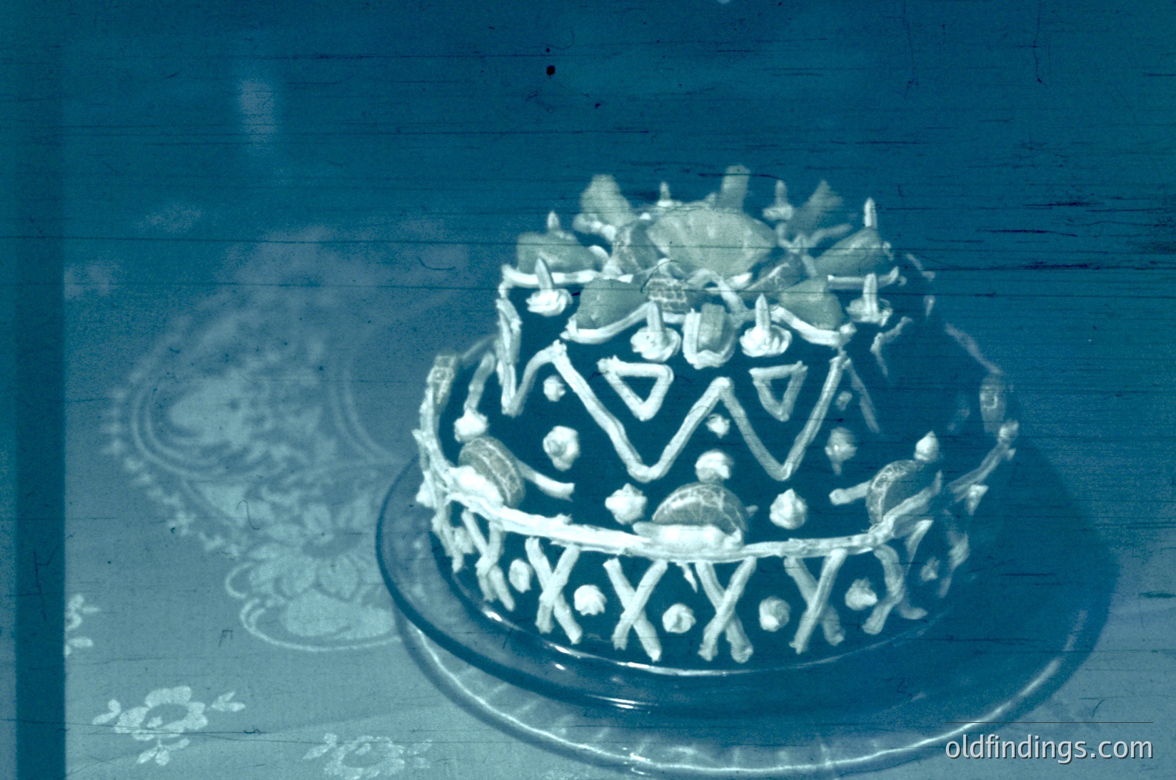 Vintage sepia-toned cake adorned with geometric icing patterns and decorative sugar accents, likely a wedding or ceremonial dessert. Blue-tinted filter suggests mid-20th-century photography. Intricate floral motifs on the tablecloth hint at formal occasion. Ideal for historical research on wedding traditions or retro dessert design.