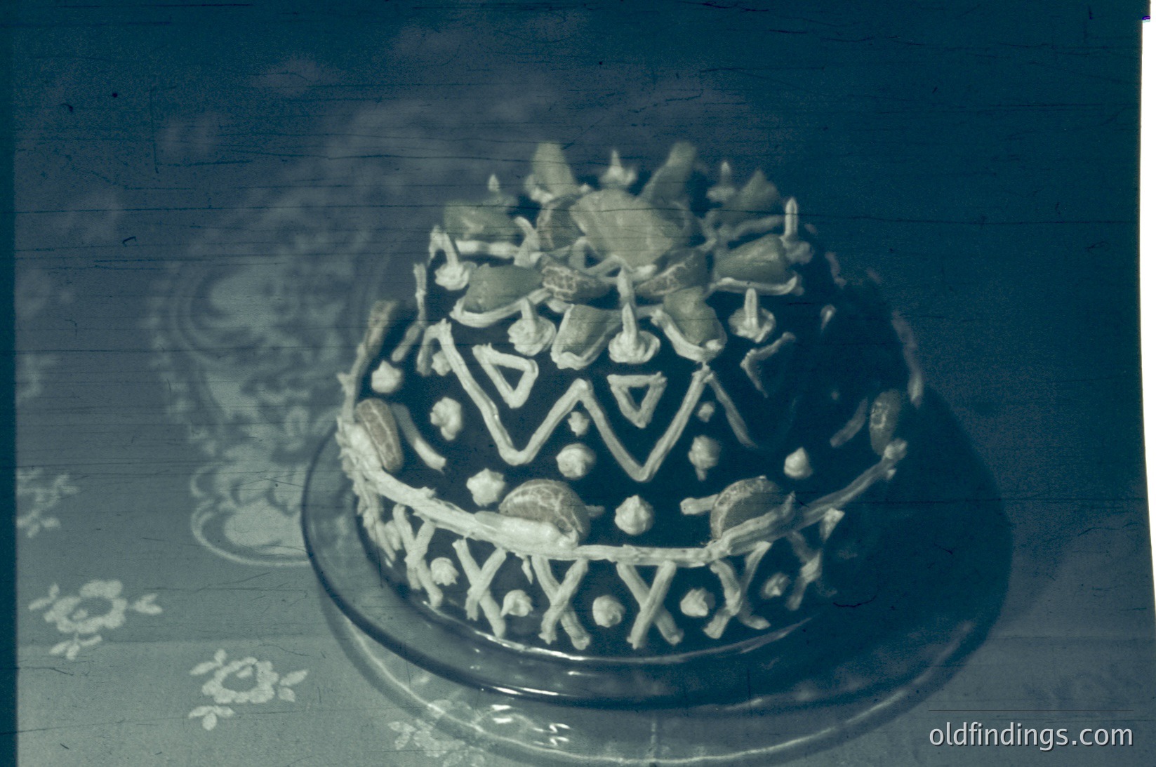 Vintage decorative cake topper featuring geometric and floral motifs, likely from the 1950s–1970s. Intricate silver filigree and rhinestone accents suggest use for weddings or formal celebrations. Background floral pattern hints at mid-century home decor.