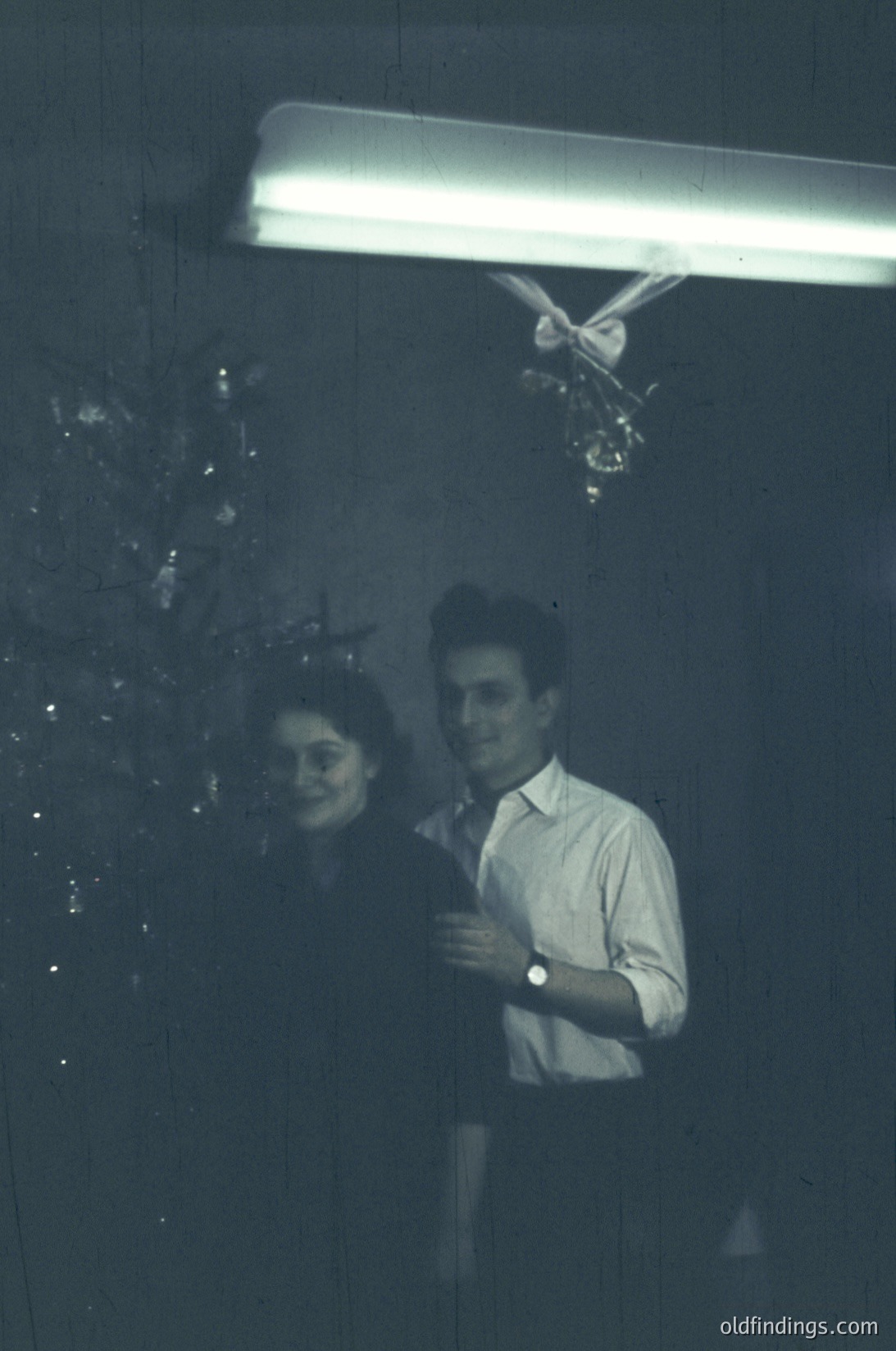 Black-and-white candid shot of two people standing under a fluorescent light, framed by a curtain of hanging ornaments. The man wears a short-sleeve button-down shirt; the woman’s face is partially obscured by the light. Likely a 1960s–1970s indoor holiday scene, possibly a home or office.