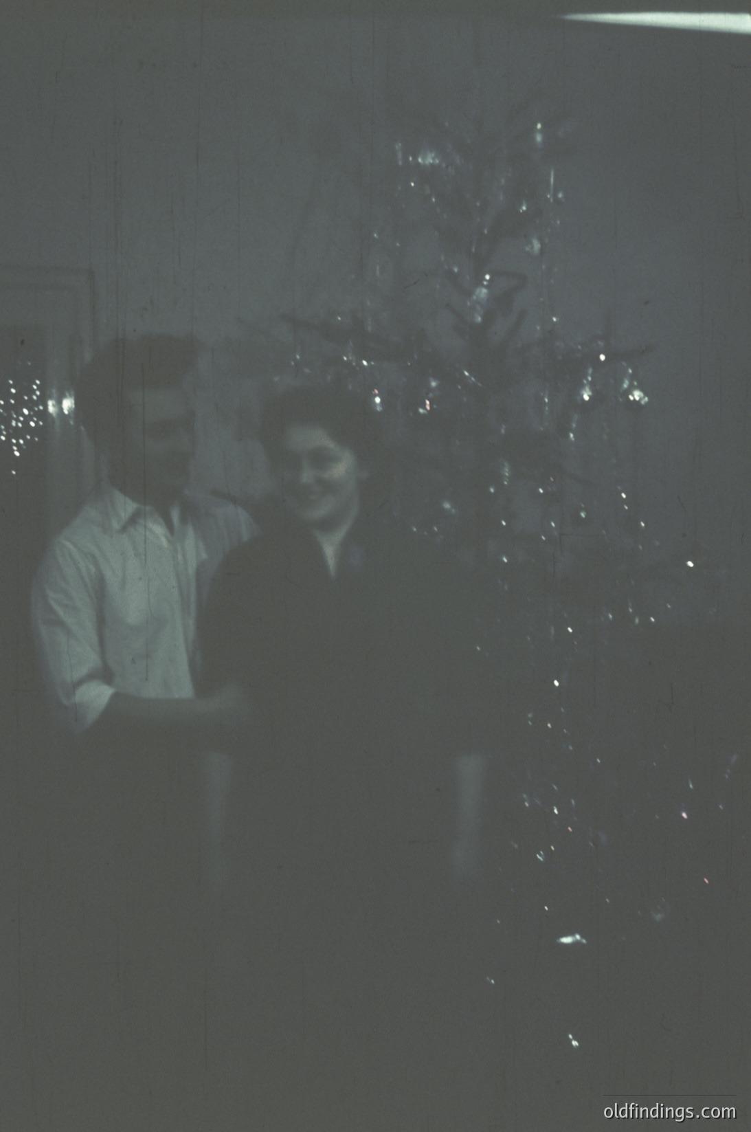 Vintage black-and-white photo of two people posing beside a decorated Christmas tree in an indoor setting, likely a home or small gathering. The man wears a button-down shirt, the woman a dark top with a light collar. Tree adorned with string lights and ornaments. Style suggests mid-20th century (1950s–1960s).