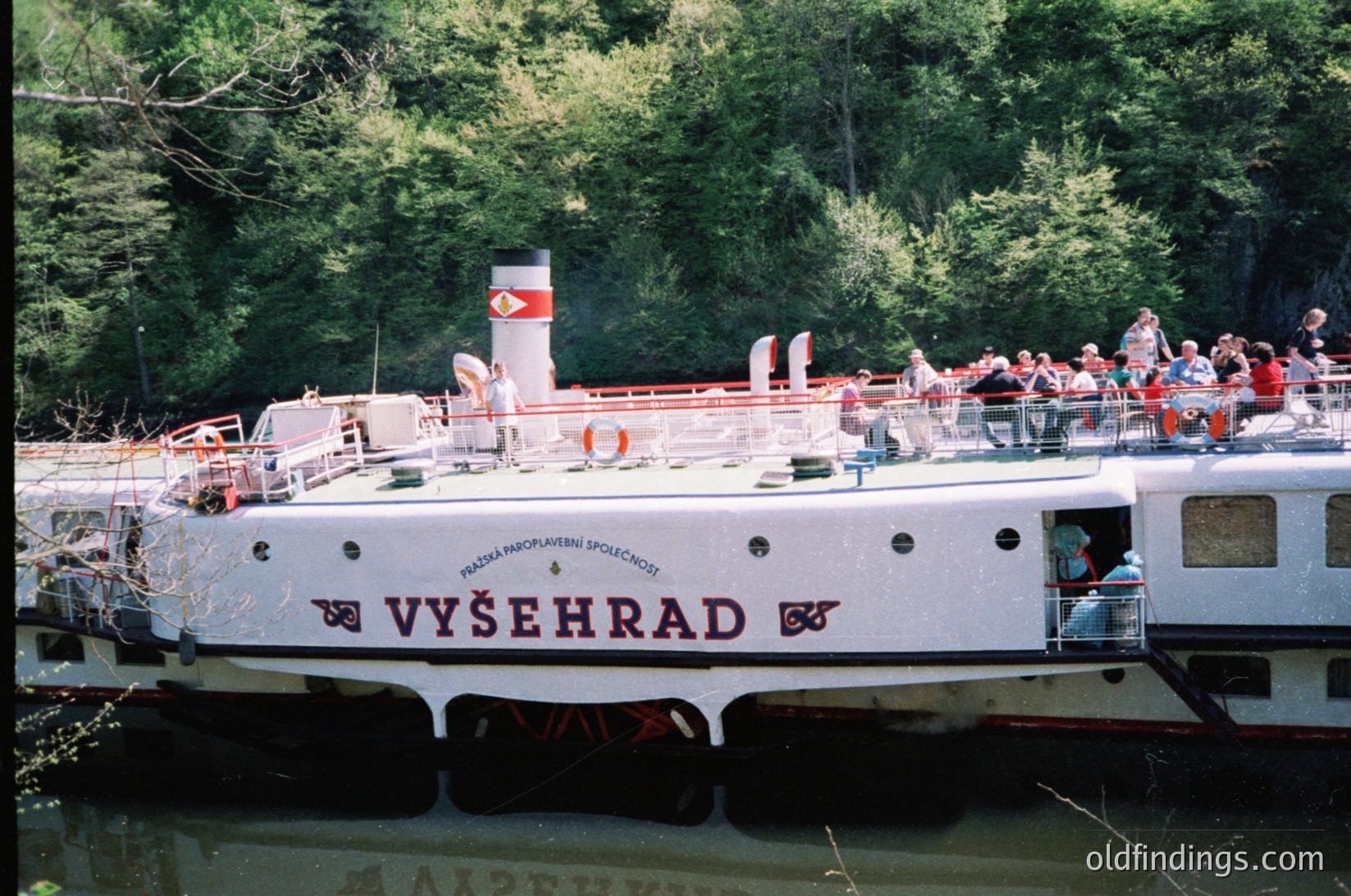 Vintage passenger ferry "Vyšehrad" on the Vltava River, Prague, Czech Republic. White hull with red accents, carrying tourists along a wooded riverside. šehradFerry --- *Note: The approximate time period is inferred from the ferry’s design and passenger attire, suggesting the 1980s. The "Praha" text confirms the Czech Republic.*
