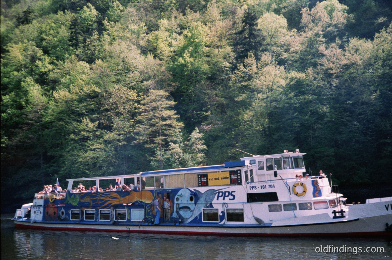 Vintage passenger ferry on a serene river, surrounded by dense forest. The boat features colorful murals of fish and maritime symbols, with "PPS" and "PP-181 704" markings. Mid-20th century design, likely Eastern European.