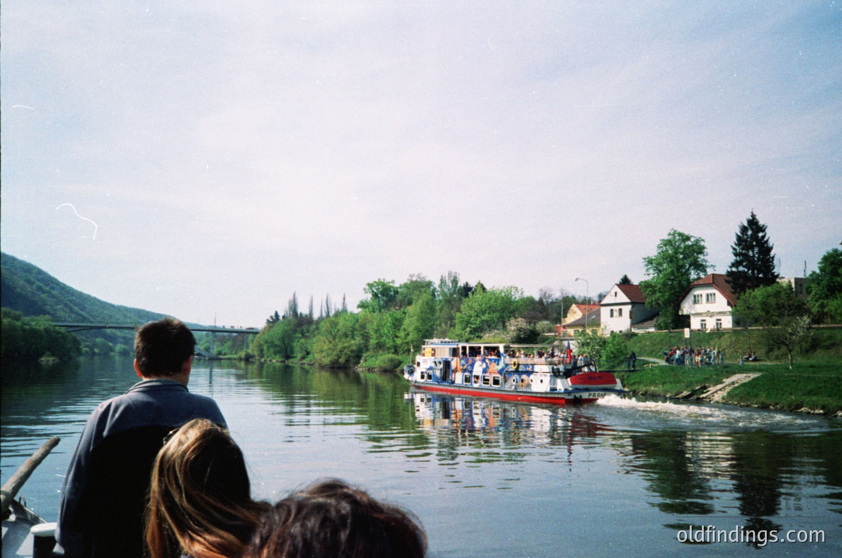 A vintage river scene featuring a passenger ferry with red and white hull, carrying people on a calm waterway. In foreground, two individuals observe from a smaller boat. Lush greenery and modest houses line the riverbank, with distant hills under clear skies. Likely Eastern Europe, mid-20th century.