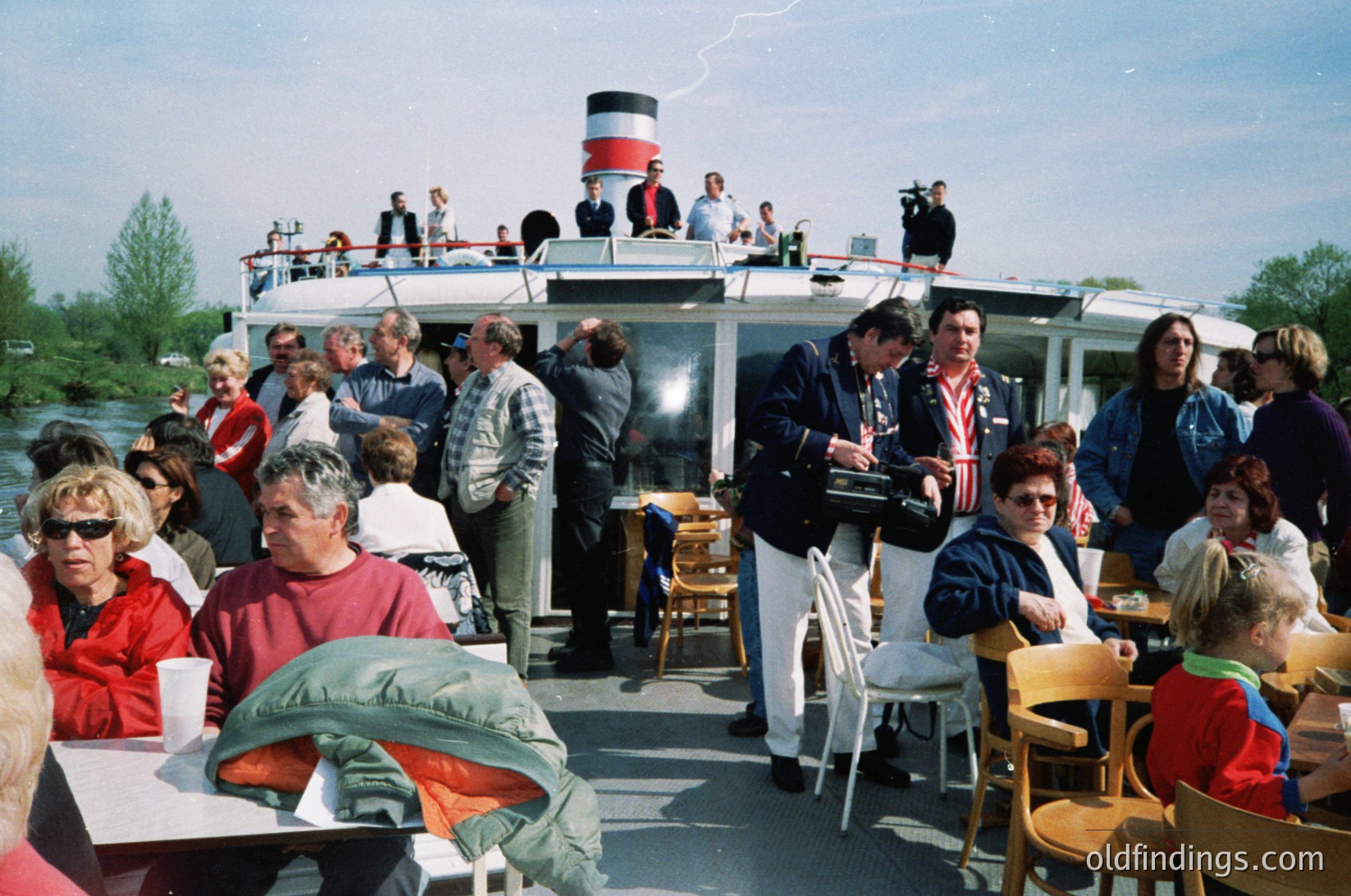 Crowded deck scene aboard a retro riverboat with red-and-white funnel, likely 1980s–1990s. Patrons in casual attire—jackets, sunglasses, and summer clothes—dine at tables while others stand near the railing. Lush greenery and water in background suggests a European river cruise. Ideal for vintage travel, tourism, and nostalgic stock imagery.