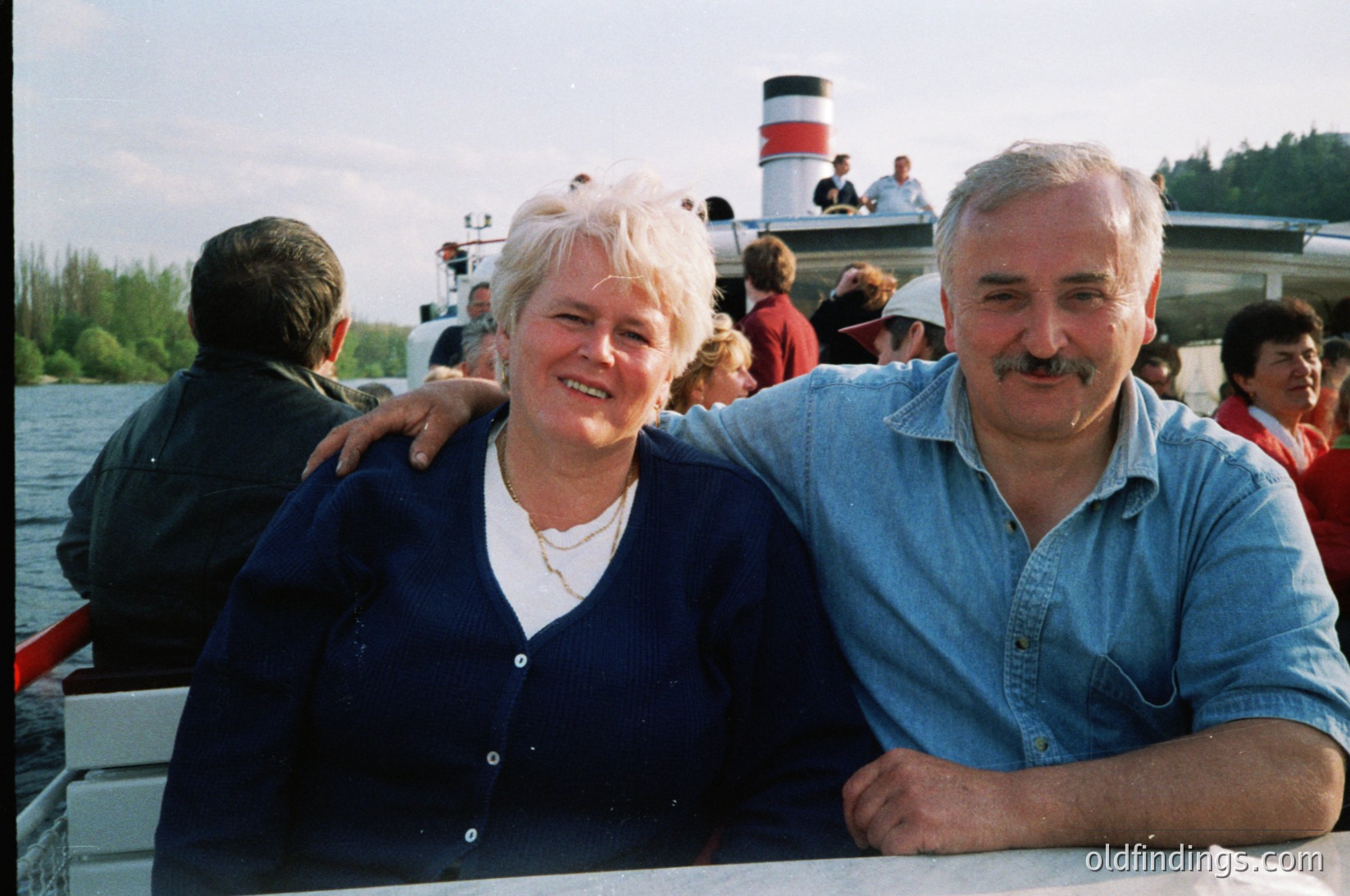 Two adults pose joyfully on a vintage ferryboat, arms around each other. The woman wears a dark blue cardigan over a white blouse; the man sports a light blue button-up with a mustache. Background shows passengers on deck and a red-and-white striped ferry tower. Likely late 20th century, possibly , in a coastal or lakeside setting.