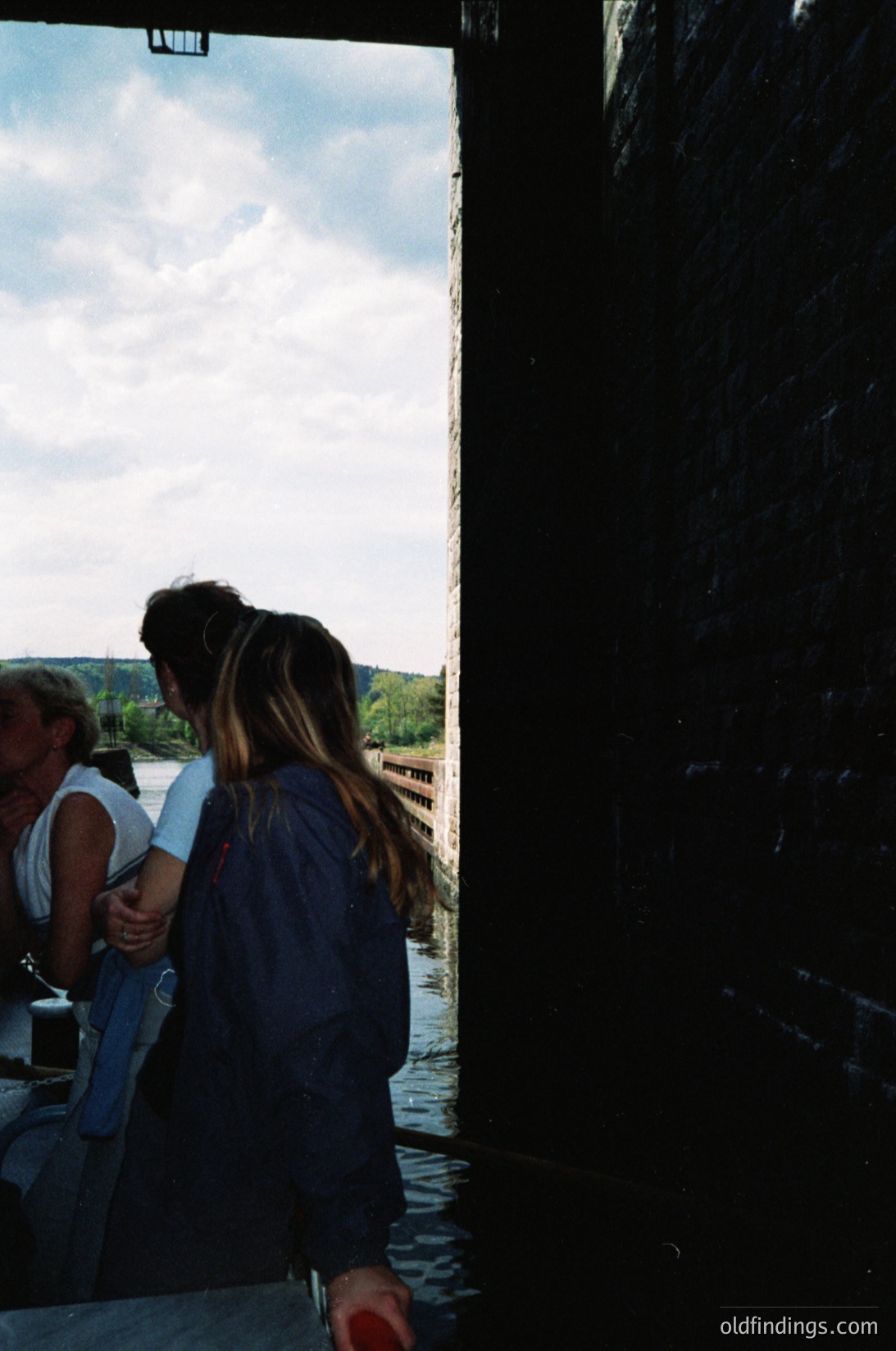 Two individuals stand indoors near a large wooden door, framed by natural light. One wears a light tank top and jeans, the other a dark long-sleeve shirt. Outside, a serene lake and forested hills stretch under a partly cloudy sky. Mid-20th century interior design with wooden paneling visible.