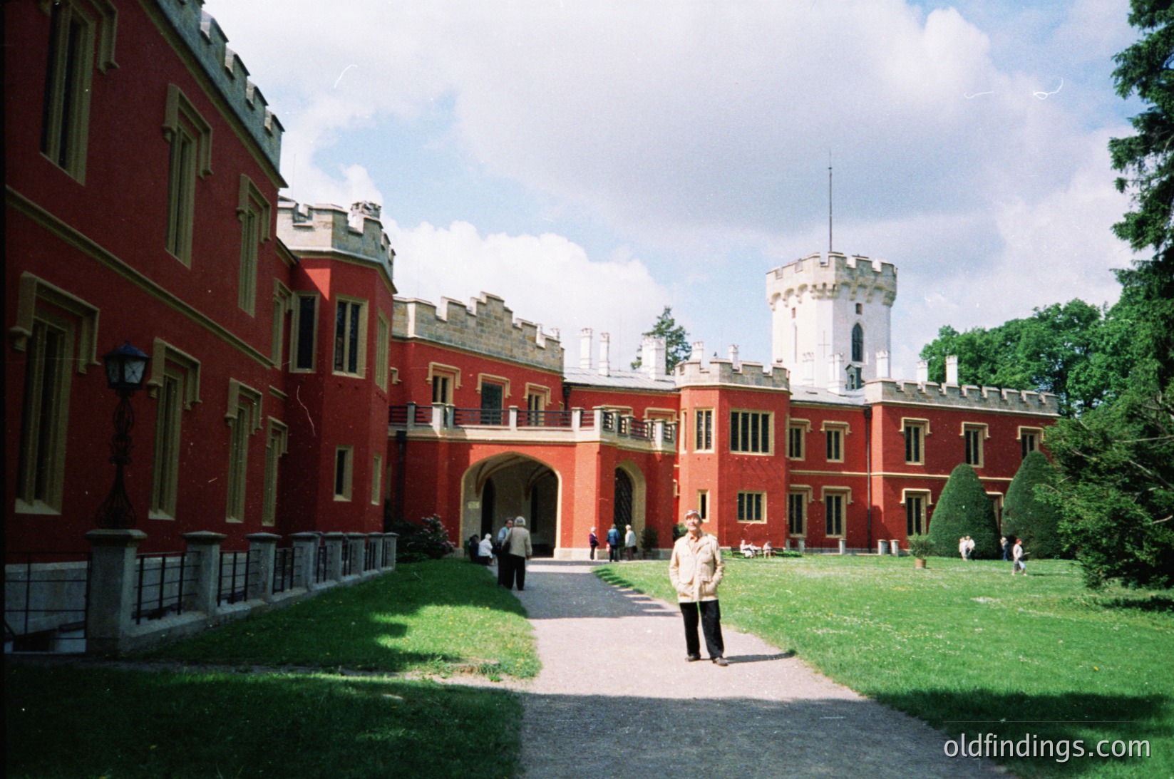 Neoclassical-style red-brick building with white accents, featuring a central archway and corner towers. Courtyard with manicured lawn, gravel path, and vintage lampposts. Two individuals in mid-20th-century attire (one in light jacket, one in dark pants) walking toward entrance.