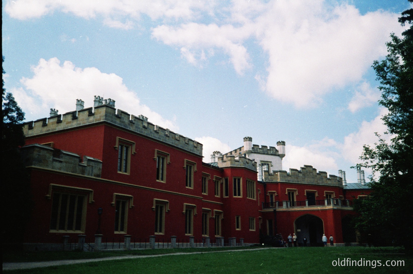 Neoclassical-style red-brick building with crenellated roofline, likely a historic estate or government complex. Symmetrical facade features tall, narrow windows and a central arched entrance. Lush green lawn and mature trees frame the scene. Potential Eastern European architecture, mid-20th century.