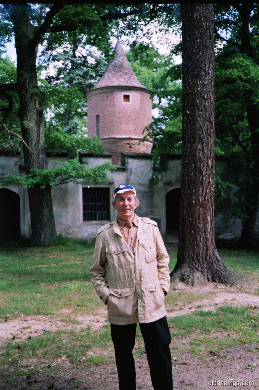 Man in vintage military-style jacket poses in front of a historic tower with conical roof and arched stone walls, surrounded by mature trees. Likely Eastern Bloc-era architecture, mid-20th century.