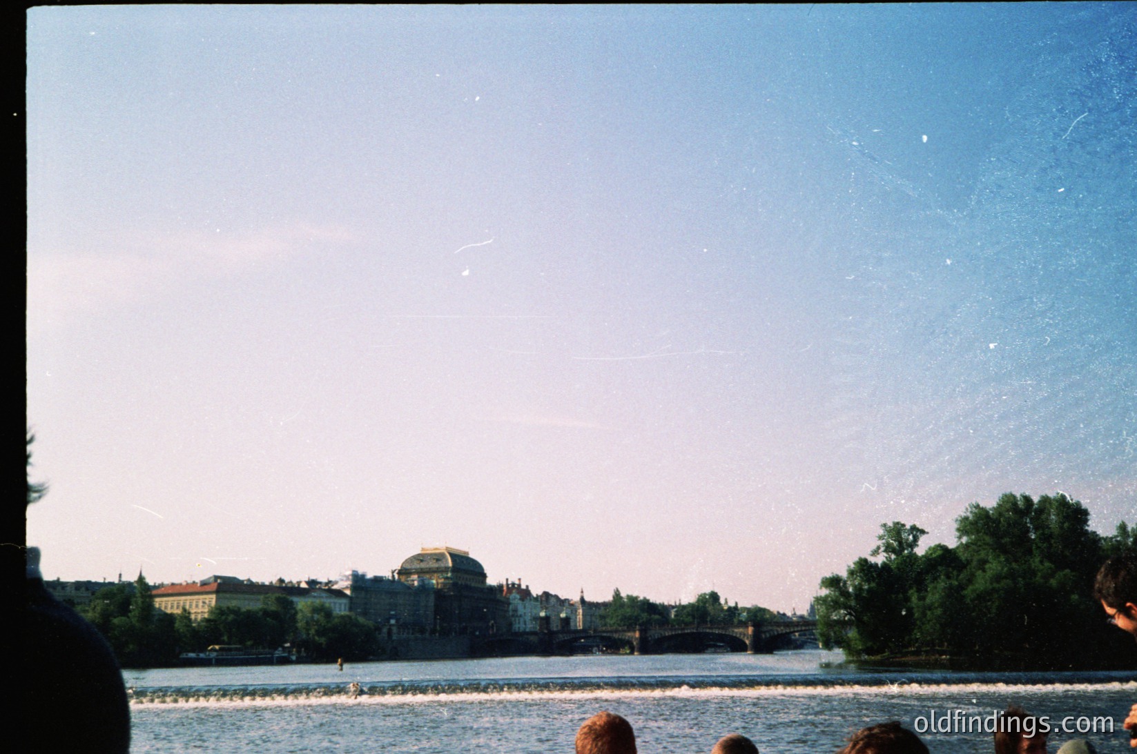 Vintage shot of a riverbank scene with a distant bridge and domed building, likely Warsaw’s Palace of Culture. Soft focus and grainy texture suggest 1960s–1980s photography. Crowd of people along the waterfront, possibly a public gathering or event.