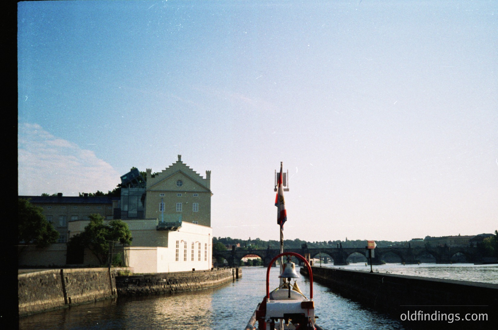 Vintage boat on calm river with classic European-style building in background, featuring pastel green walls and white trim. Bridge and distant cityscape visible across water. Likely 1980s-1990s European riverside scene.