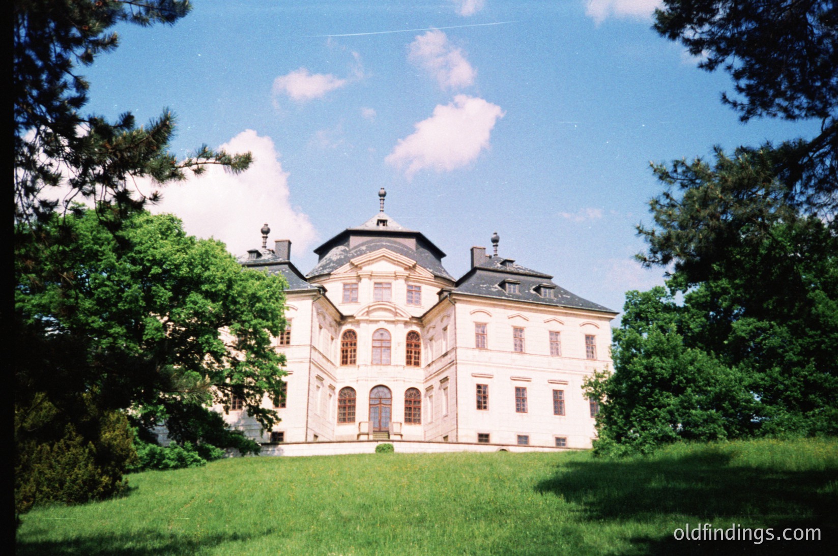 Neoclassical mansion with symmetrical façade, central pediment, and mansard roof. Lush green lawn and mature trees frame the structure, suggesting a well-maintained estate. Likely European, possibly architecture.