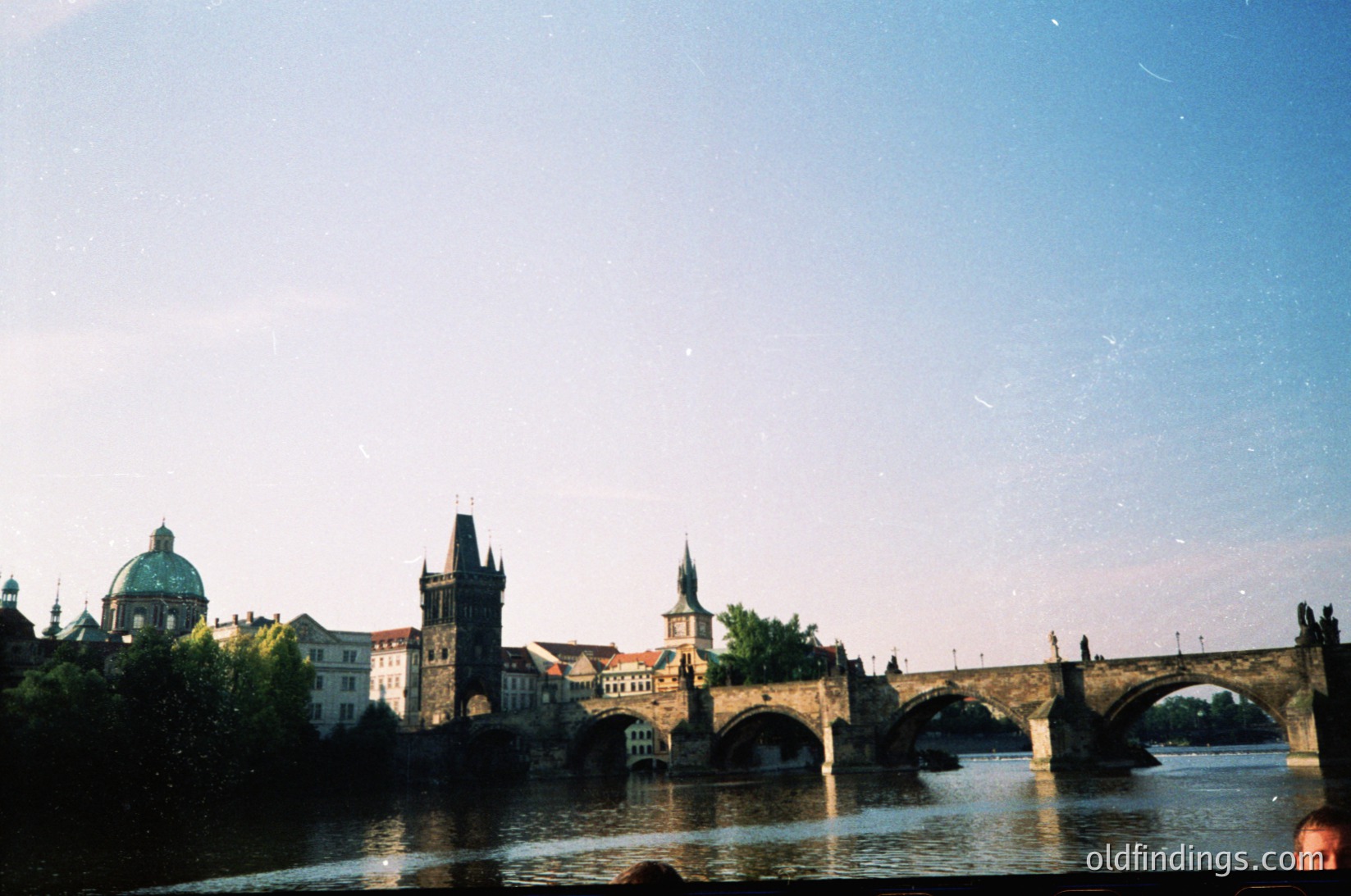 Iconic Charles Bridge spanning Prague’s Vltava River, showcasing Gothic towers and domes of historic Prague Castle in background. Mid-20th century urban landscape with vintage film grain.