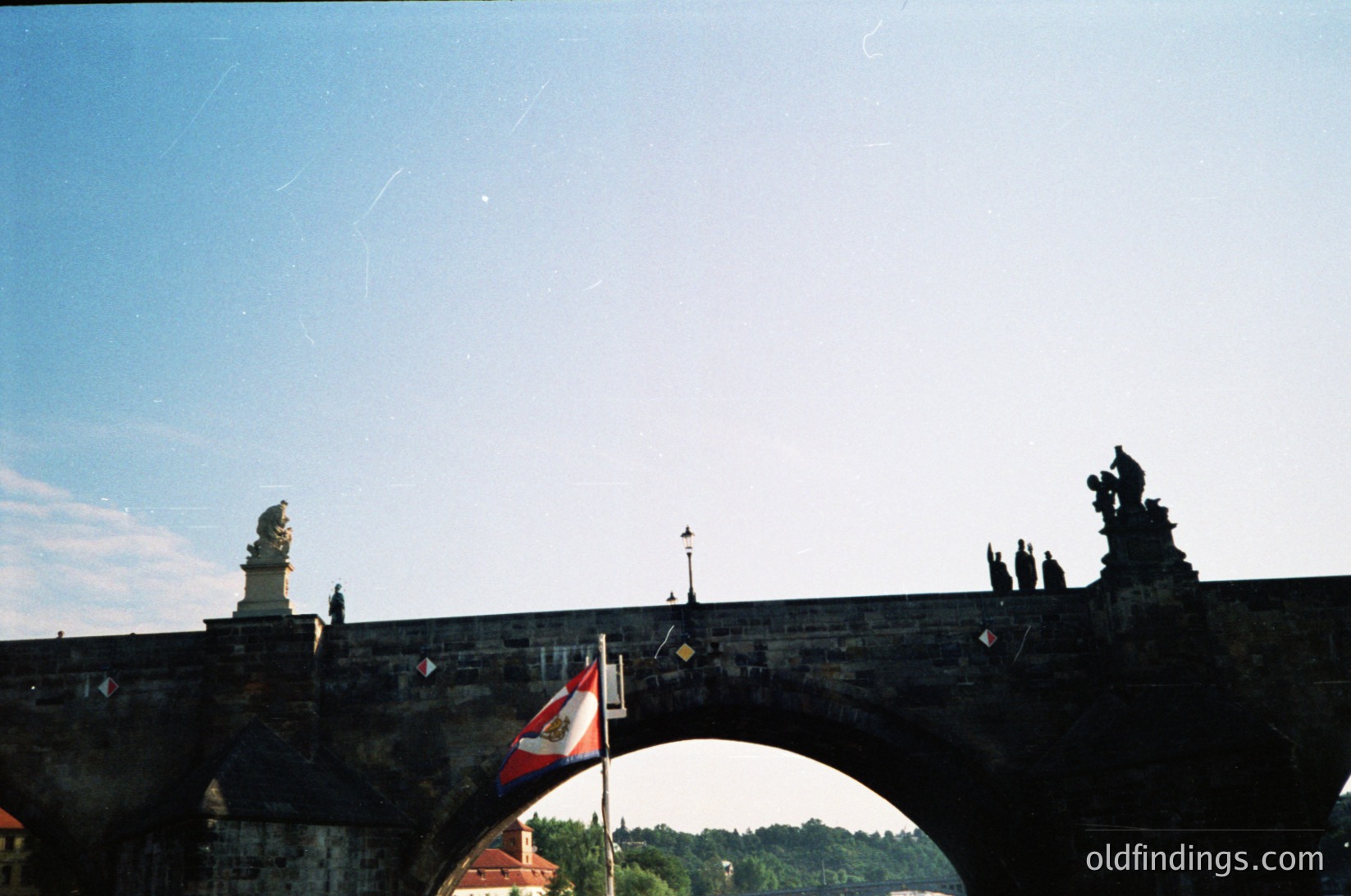 Historic stone bridge with Gothic statues and arch framing a flagpole displaying Czech and EU flags. Urban landscape with distant greenery and clear sky. Likely Prague’s Charles Bridge, 20th century.