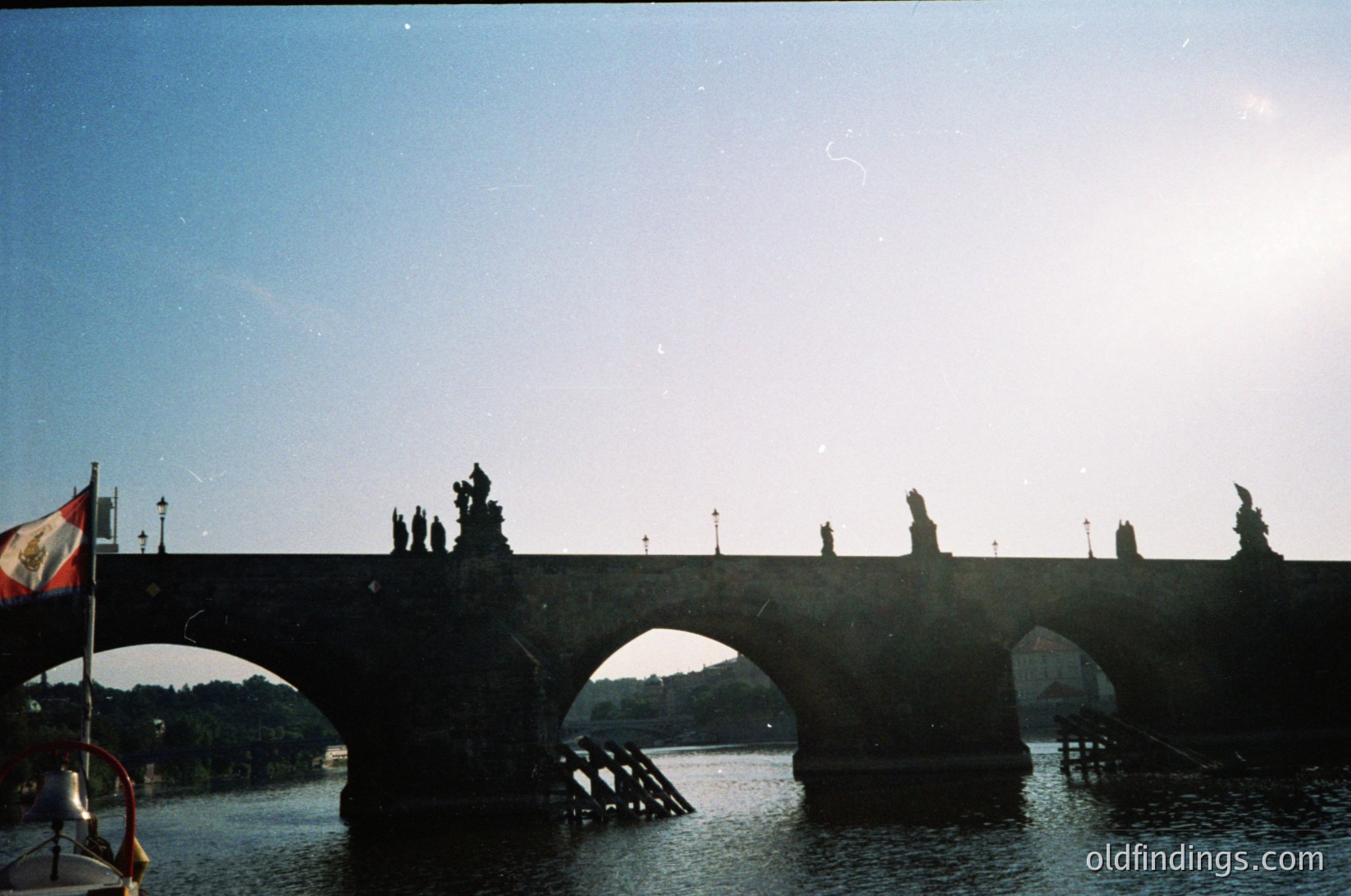 Historic stone bridge with Gothic statues over the Vltava River, likely Prague’s Charles Bridge. Sunlight casts long shadows on arches and statues. Mid-20th century (1950s–1970s) vintage filter. Iconic European architecture with religious sculptures.