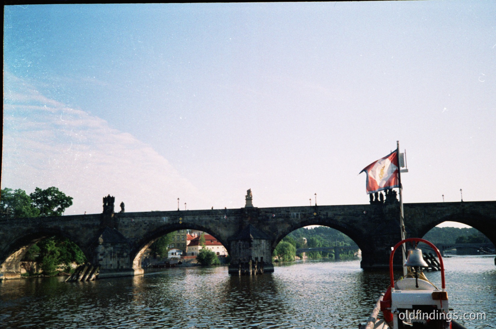 Historic stone bridge with Gothic statues spanning a river in Prague, Czech Republic. The structure features multiple arches and a prominent flagpole with a red-and-white banner. A small boat with a white cabin floats beneath. Likely or early due to film grain.
