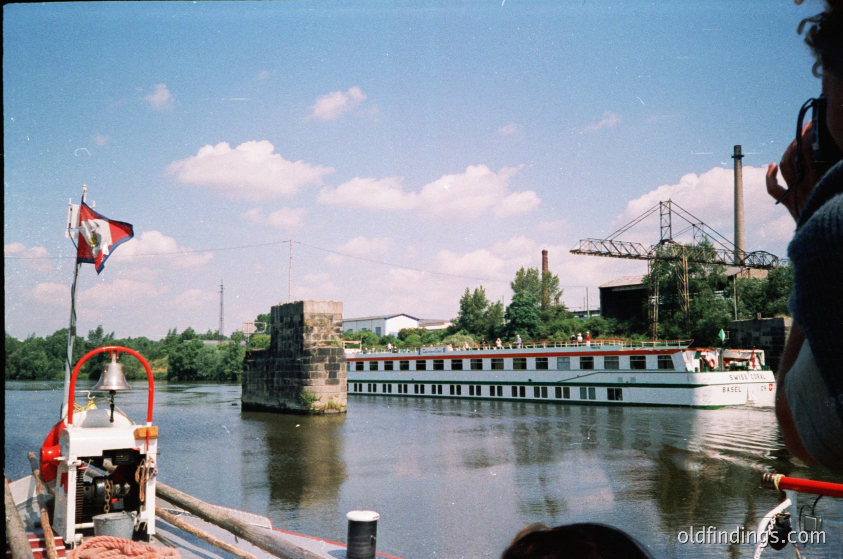 Vintage river scene featuring a mid-20th-century passenger boat with "BASIL" lettering, docked near a concrete pier. Industrial chimneys and cranes in background suggest port activity. Flag with red/yellow/blue stripes hints at Eastern European setting. Clear skies and calm water enhance nostalgic atmosphere.