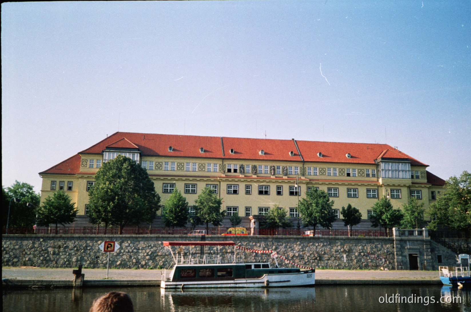 Three-story yellow building with red-tiled gable roofs, symmetrical windows, and decorative brickwork near waterfront. Stone retaining wall with small boats docked below. Lush greenery and mature trees flank the structure. Likely 1960s–1980s European institutional or public facility.