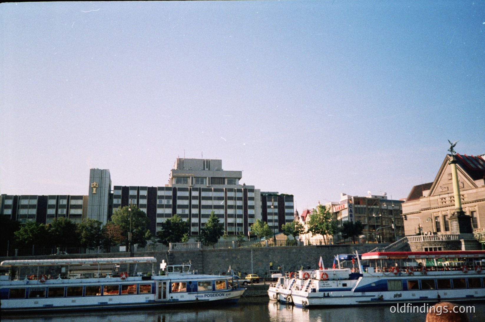 Mid-20th century riverside urban scene featuring Brutalist architecture. Prominent concrete high-rise with geometric windows dominates skyline. Foreground showcases docked passenger boats labeled "Poseidon" and "Delfin," suggesting a ferry or tour operation. Historic stone building with ornate facade and statue on roof hints at cultural significance. Clear skies and calm water indicate a serene setting. Likely Eastern European city, possibly .