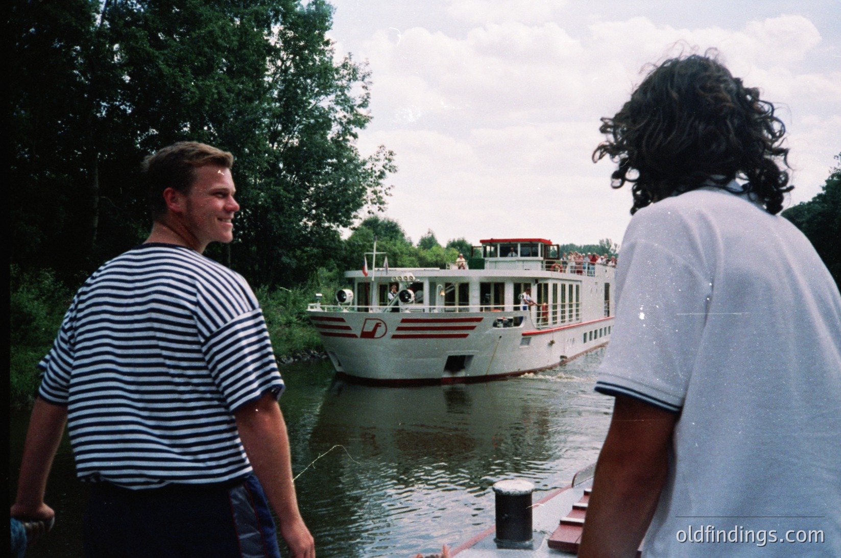 Two men stand on a dock beside a river, observing a white passenger boat with red accents and a flag. The man on the left wears a striped sailor-style shirt; the other wears a white T-shirt. Lush greenery and trees frame the scene, suggesting a serene riverside location. Likely late 20th century, possibly .