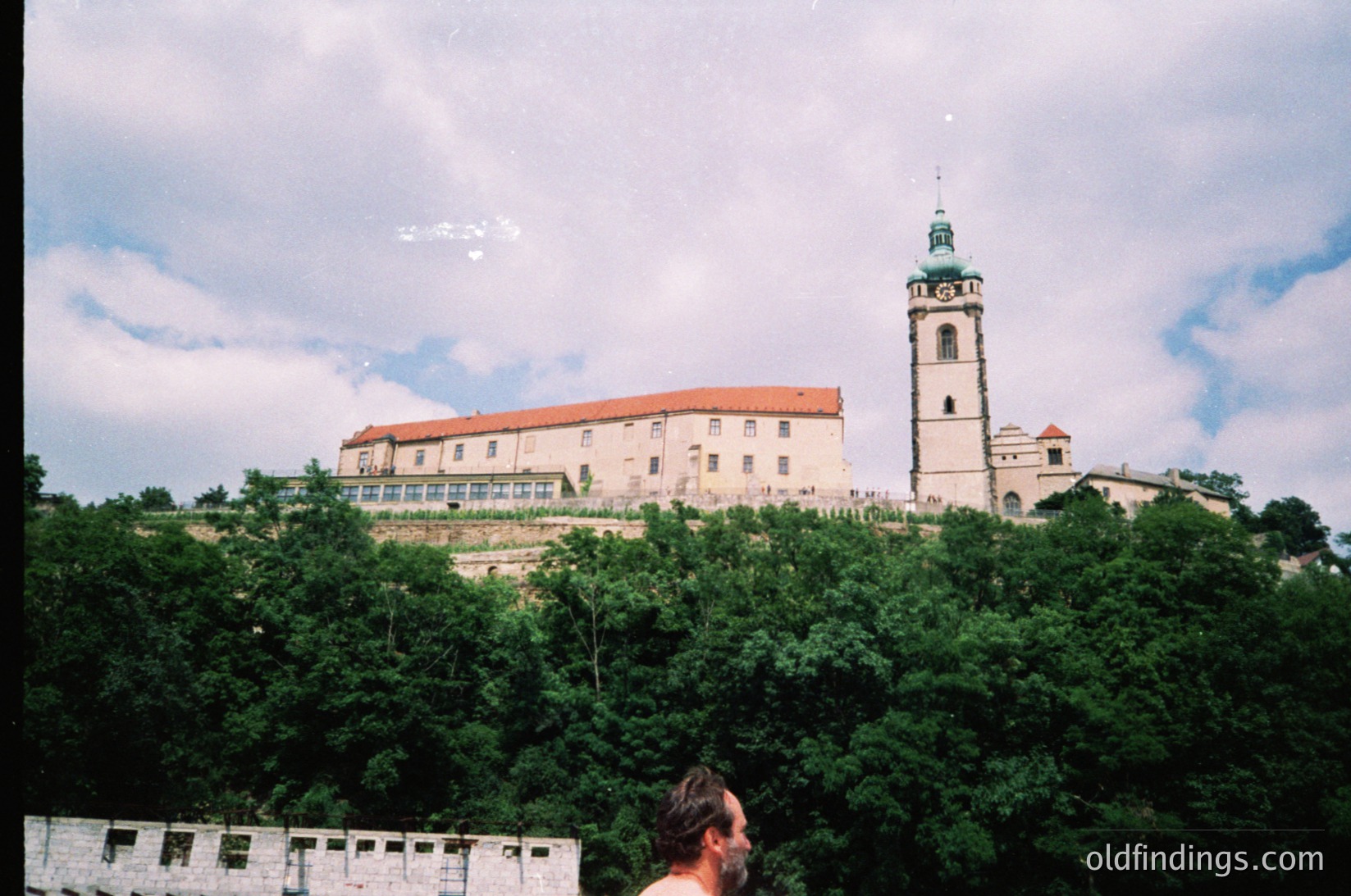 Historic fortress complex perched atop a hill, featuring a prominent bell tower and red-tiled rooftops. Lush greenery surrounds the base, blending into a stone wall at the foreground. Likely Eastern European, possibly architecture.