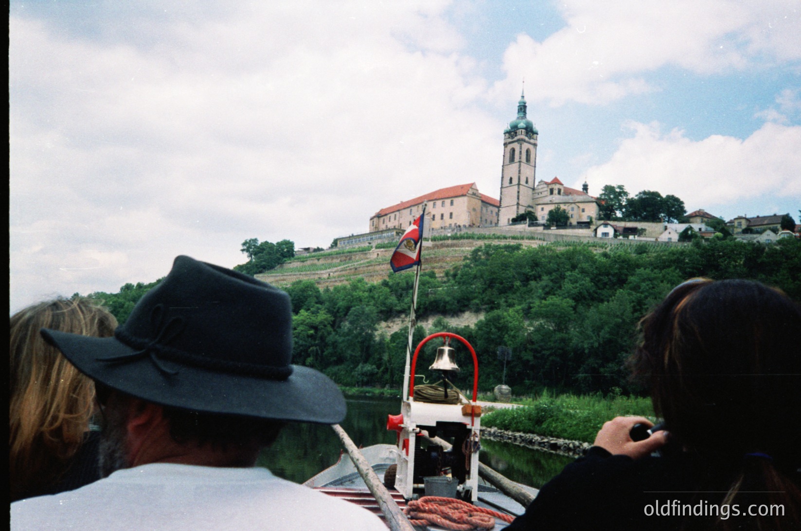 Boat tour view of medieval fortress with bell tower atop hillside, featuring red-and-white flag. Foreground shows passengers in casual summer attire, including wide-brimmed hats. Likely
