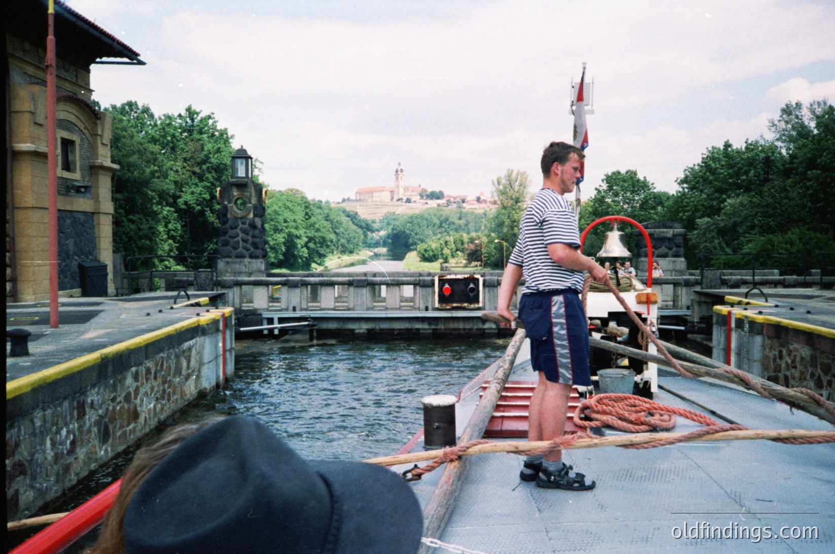 Lockkeeper operating a canal lift bridge, wearing striped shirt and holding a rope bell. Historic European town with castle ruins in background. Mid-20th century industrial waterway scene.