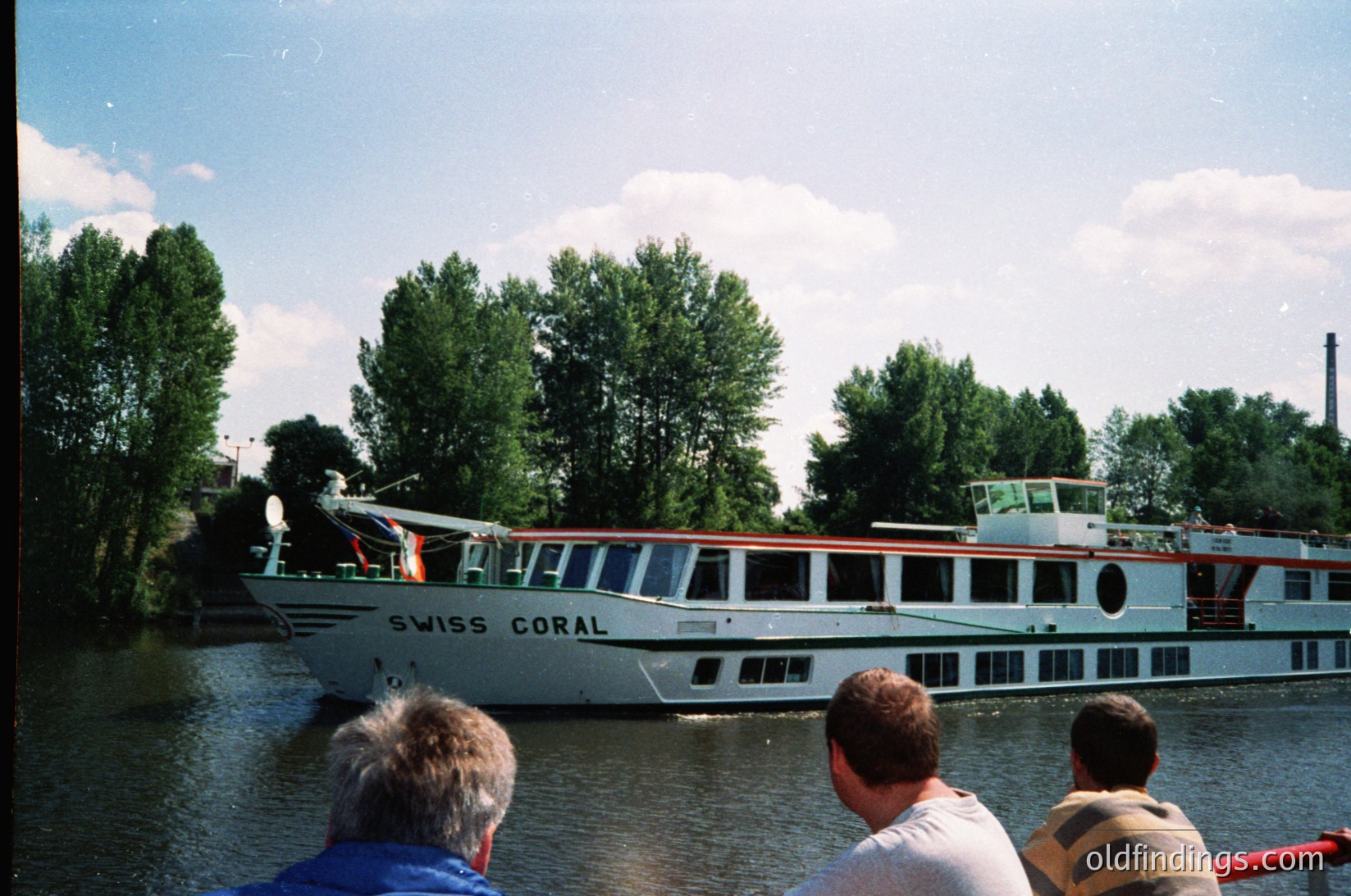 A vintage river cruise ship, *Swiss Coral*, glides along a calm waterway surrounded by lush greenery. The vessel’s classic design features white hulls with red accents and multiple decks. Passengers on a smaller boat in the foreground observe the scene. Likely taken in the 1980s–1990s in Europe.