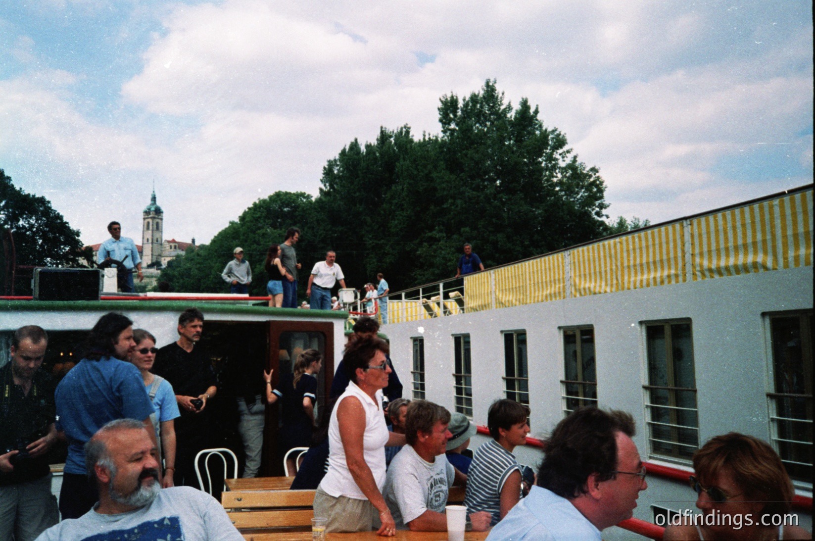 Vintage outdoor gathering on a floating restaurant or event venue, likely from the 1980s–1990s. Gray concrete structure with yellow striped awnings, surrounded by lush greenery. Crowd of casually dressed adults and children socializing, some seated at tables with drinks. Distant church tower visible, suggesting urban waterfront setting. Ideal for nostalgic or historical research on social dining trends.